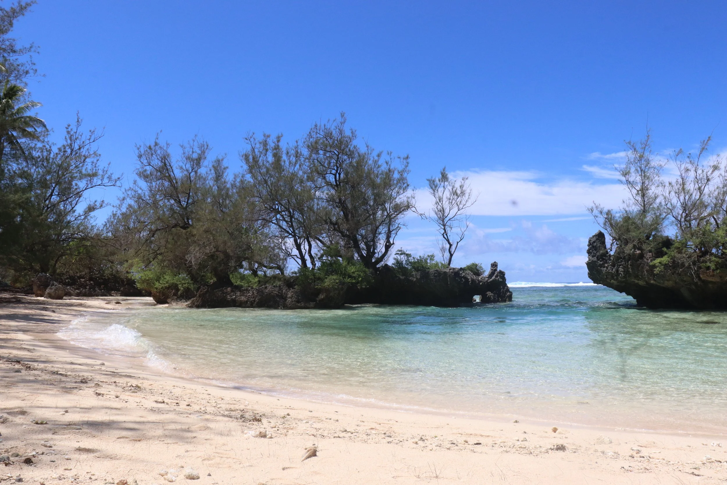 Une plage de sable blanc avec des eaux turquoise, entourée d'arbres et de rochers, sous un ciel bleu avec quelques nuages.