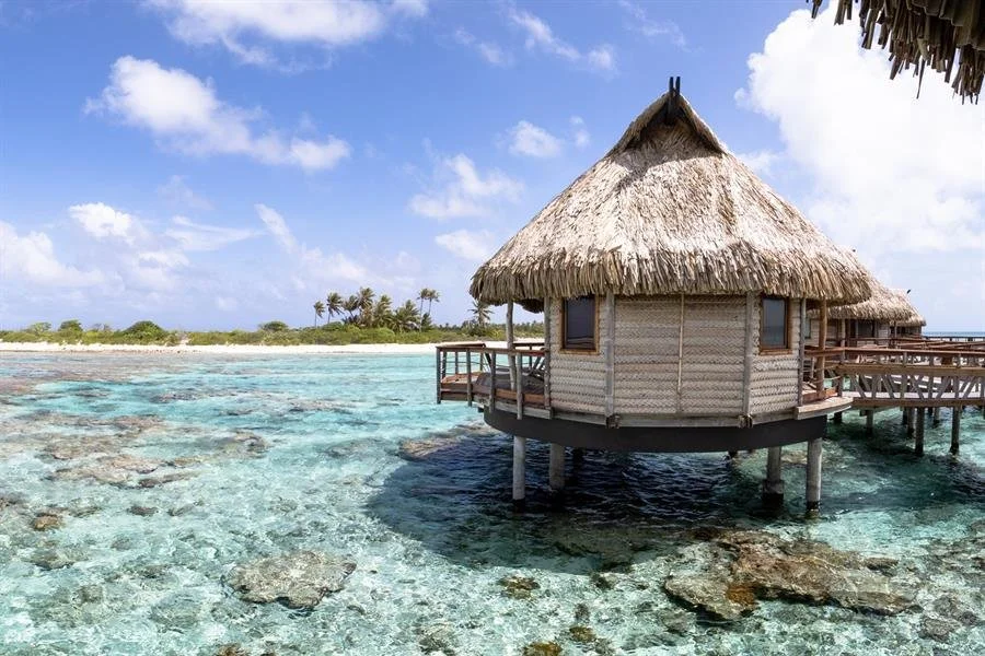 Bungalow en bois sur pilotis au-dessus de l'eau turquoise claire, avec une plage de sable fin et des palmiers au loin, sous un ciel bleu avec quelques nuages.