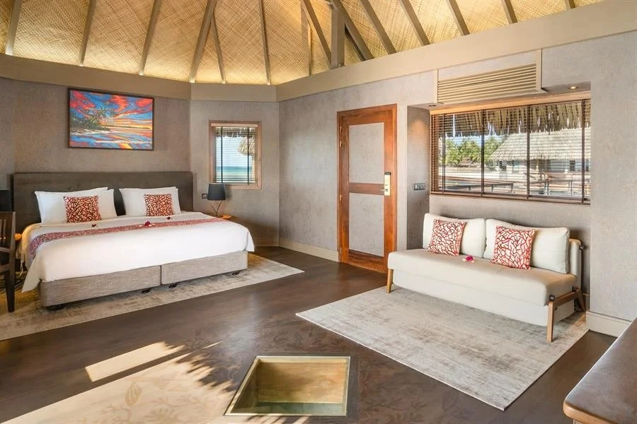 Chambre d'hôtel avec un grand lit, canapés, fenêtres avec vue sur des bâtiments et des arbres, décoration moderne, plafond en bois