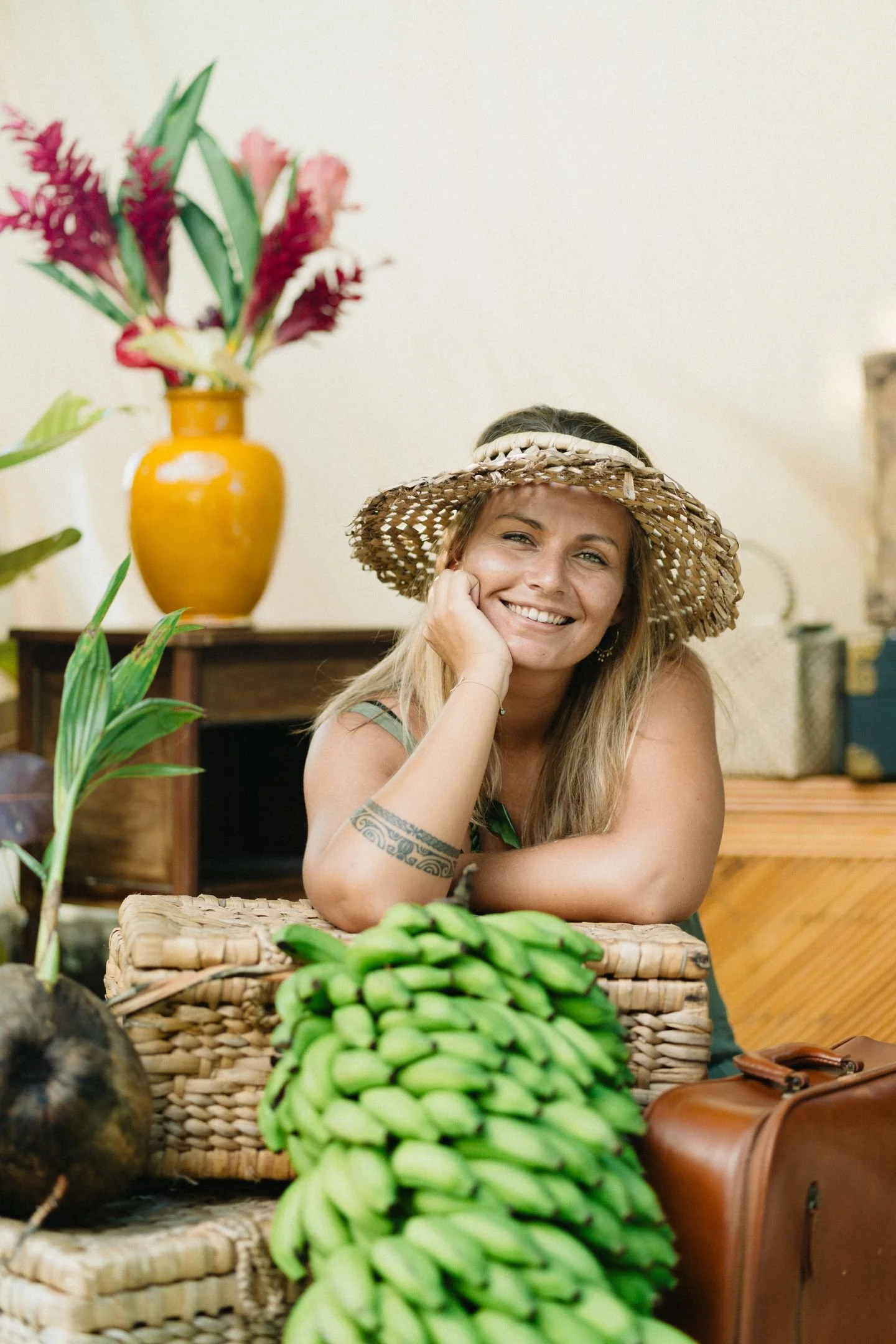 Femme souriante portant un chapeau en paille, assise à une table avec des bananes et un panier en osier, en intérieur.