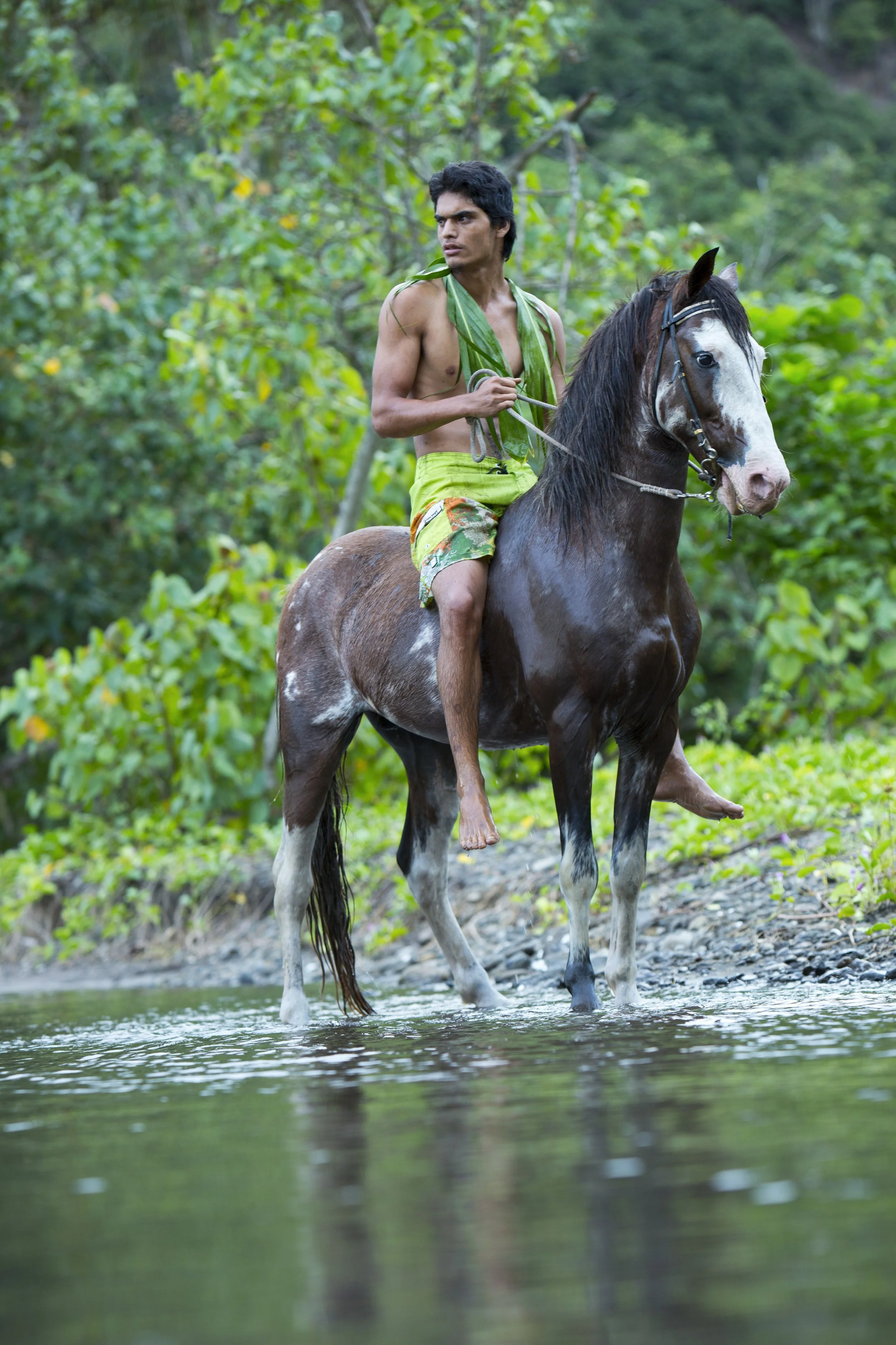 Un jeune homme à la peau foncée, sans chemise et portant un short coloré, monte à cheval dans une rivière entourée de végétation dense.