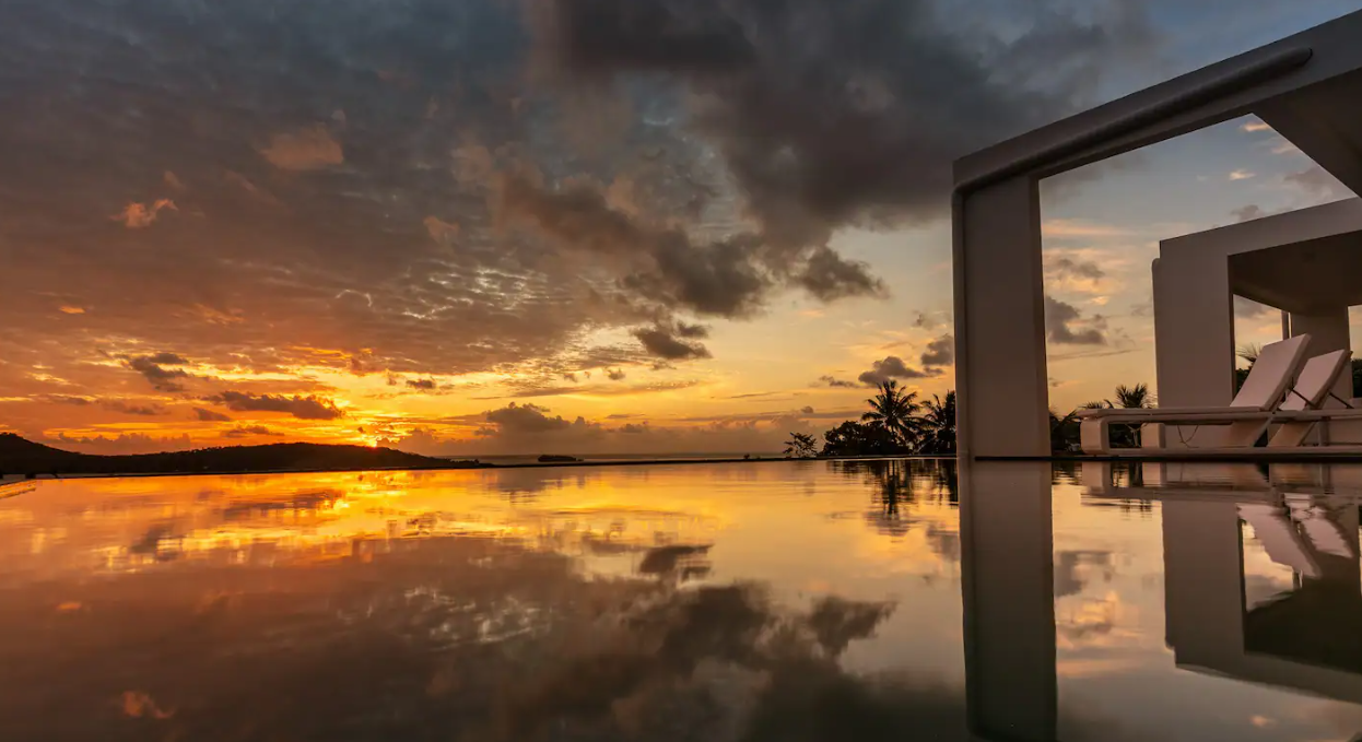 Coucher de soleil sur une piscine à débordement d'une station balnéaire avec des chaises longues et des palmiers en arrière-plan.