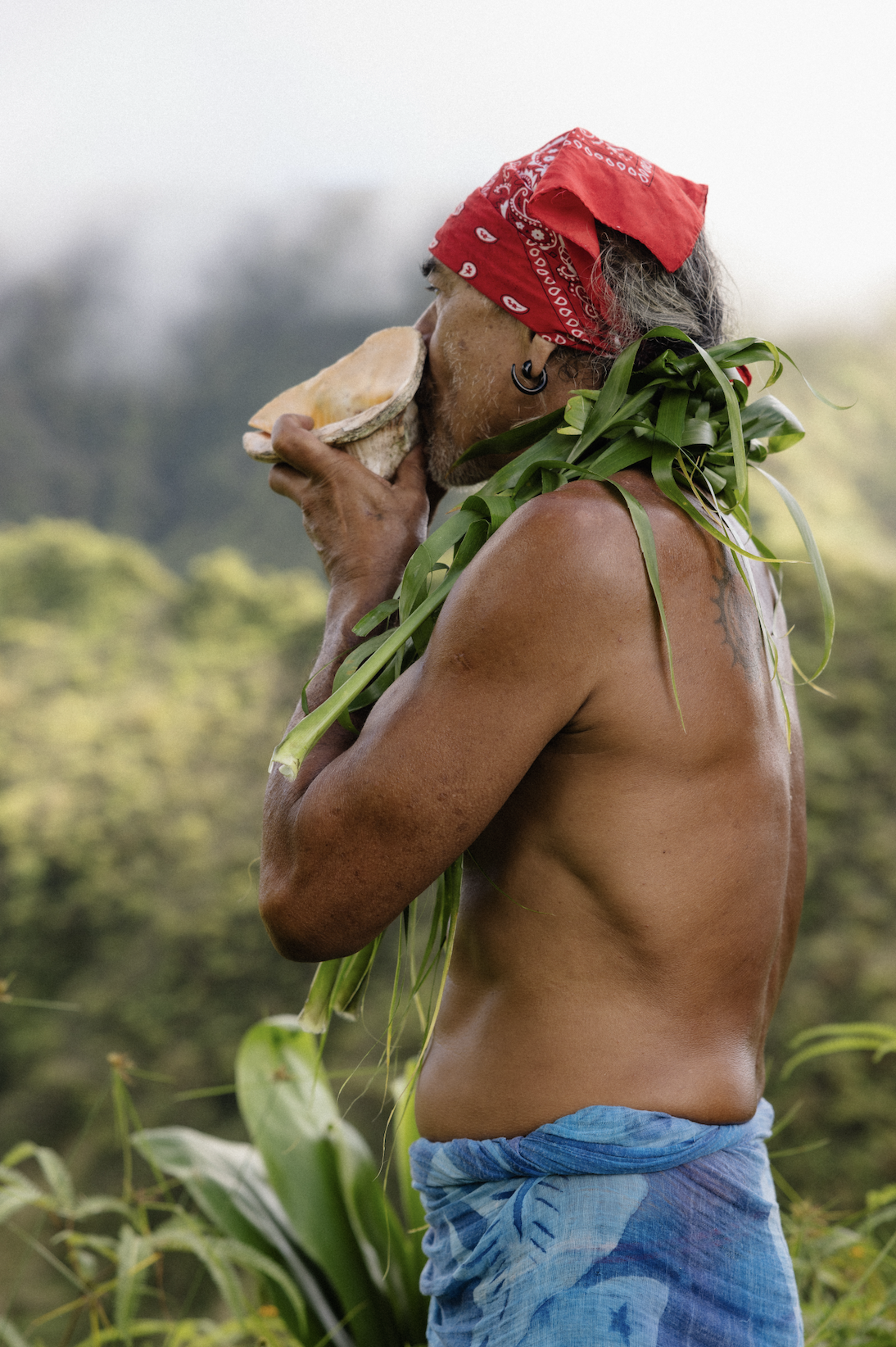 Un homme nu avec un bandana rouge sur la tête, portant des feuilles vertes sur l'épaule, joue d'un instrument en buvant de la nourriture dans un paysage naturel