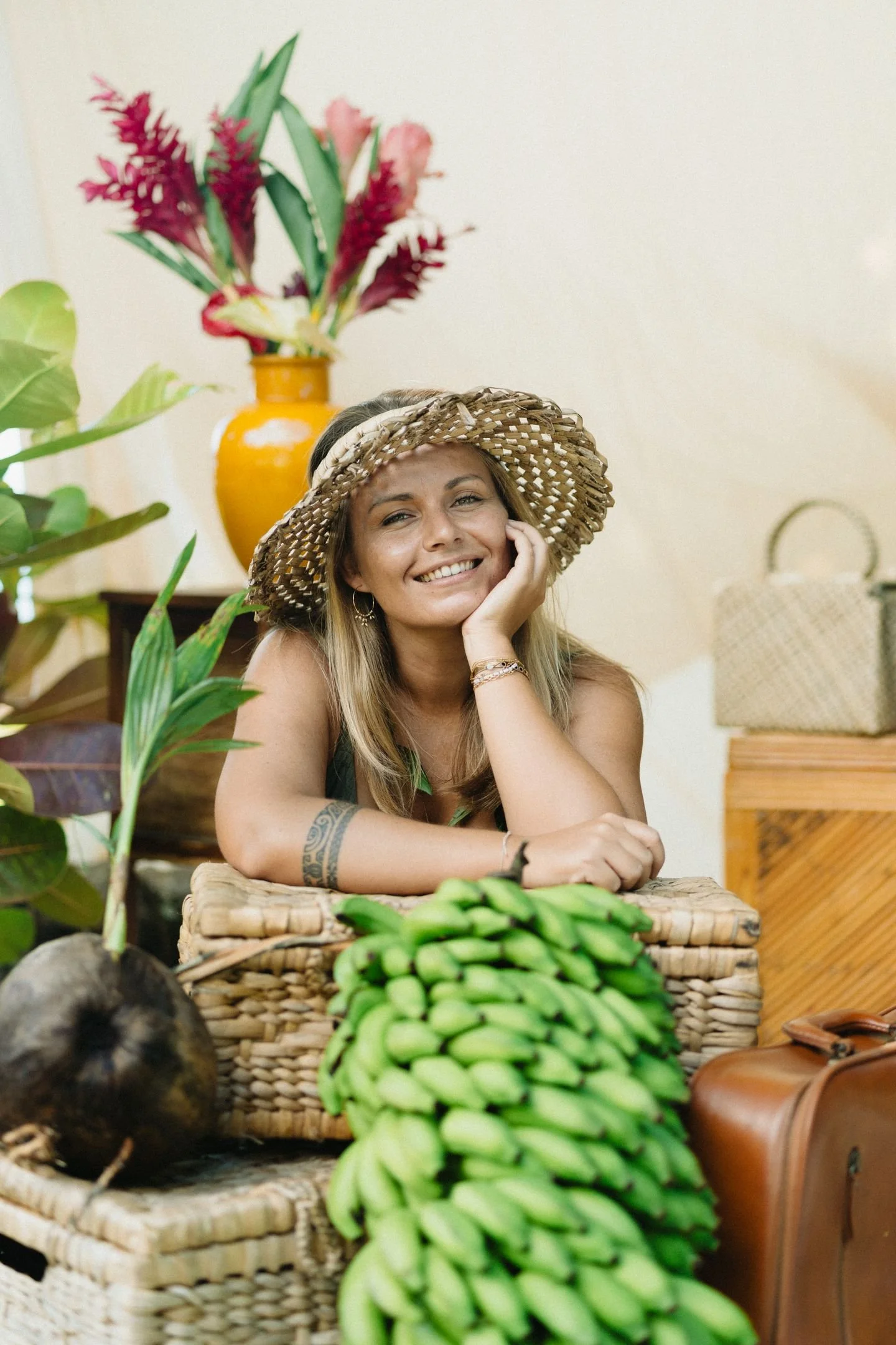 Une femme souriante portante un chapeau en paille, posant près de paniers de fruits, notamment des bananes, dans un environnement intérieur avec des plantes et une décoration rustique.