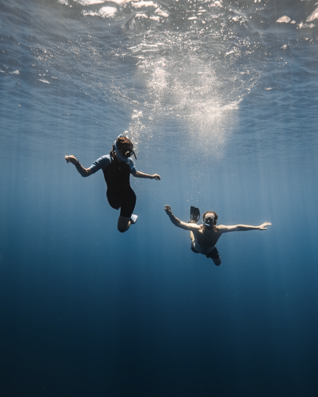 Deux personnes nageant sous l'eau, portant des lunettes de plongée, avec un fond d'eau bleue profonde.