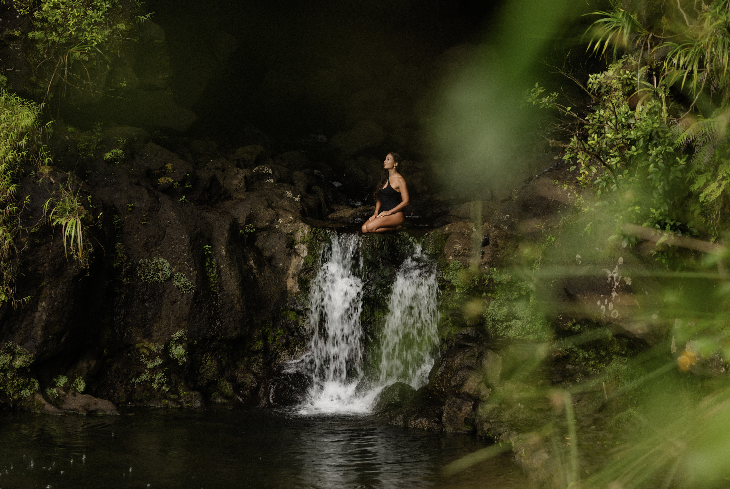 Une femme assise en meditant au centre d'une cascade entourée de végétation luxuriante dans une forêt tropicale.