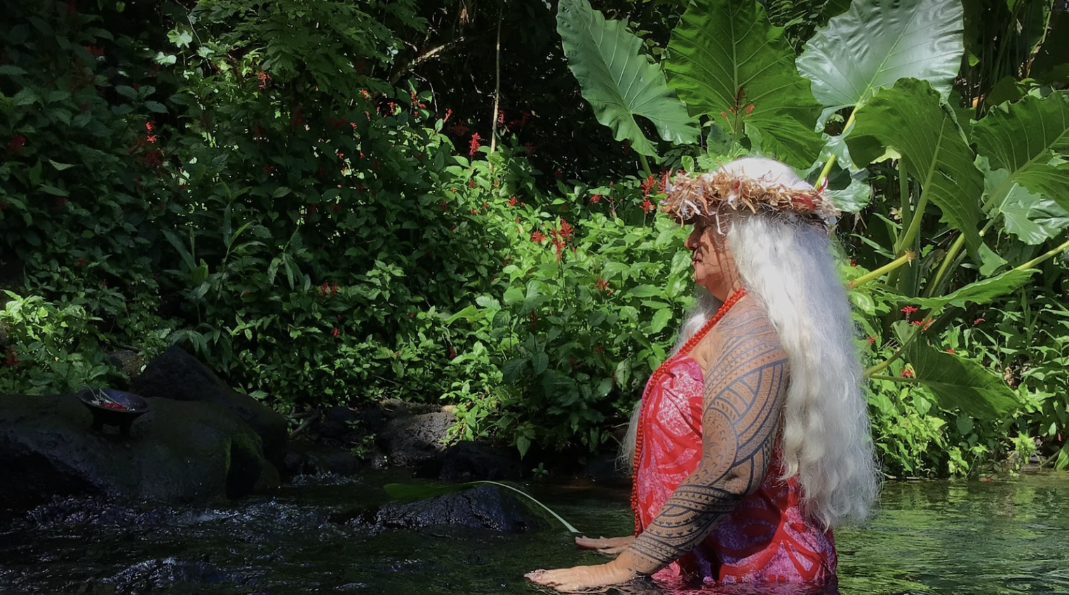 Une femme tahitienne effectuant une cérémonie dans une rivière entourée de végétation dense, portant un paréo rose, un collier rouge, un chapeau de feuilles et ayant de longs cheveux gris.