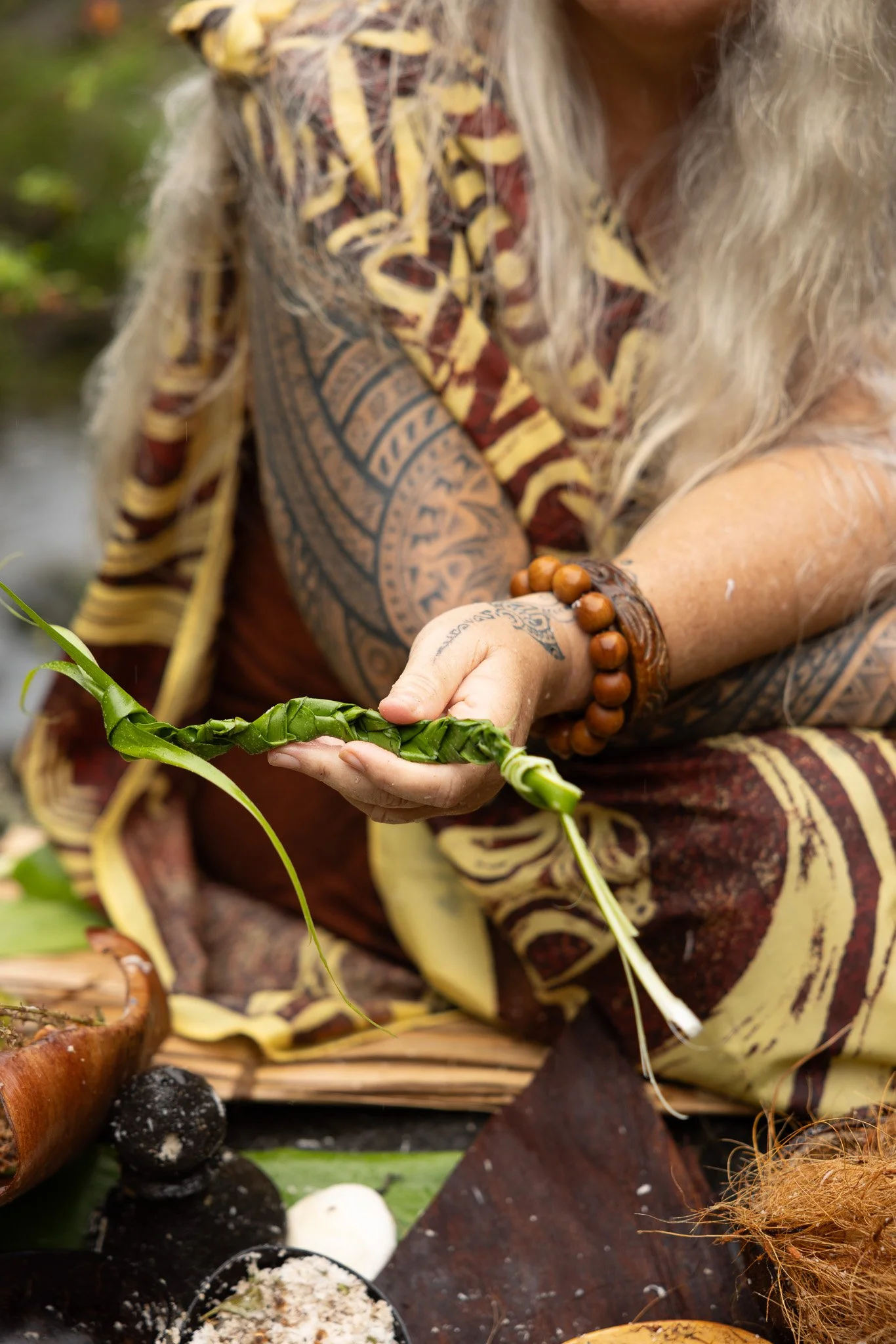 Une personne avec des tatouages et des bracelets en bois, portant une chemise à motifs, tenant une tige de bambou dans un environnement naturel.