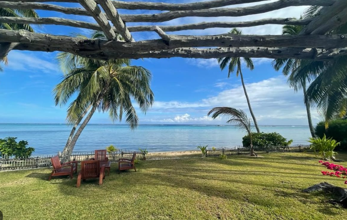 Terrasse en bois avec des chaises en bois, vue sur la mer à travers des palmiers, ciel bleu avec quelques nuages.