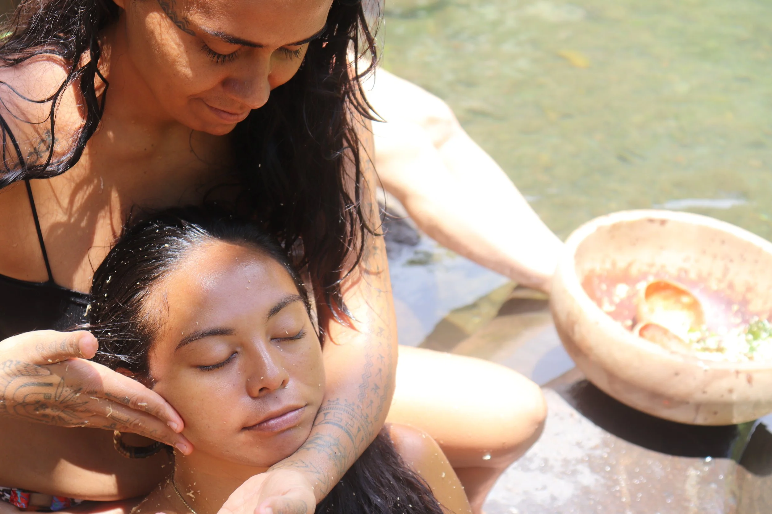 Deux femmes en train de pratiquer un massage ou une relaxation au bord de l'eau, avec un bol en pierre visible à côté.