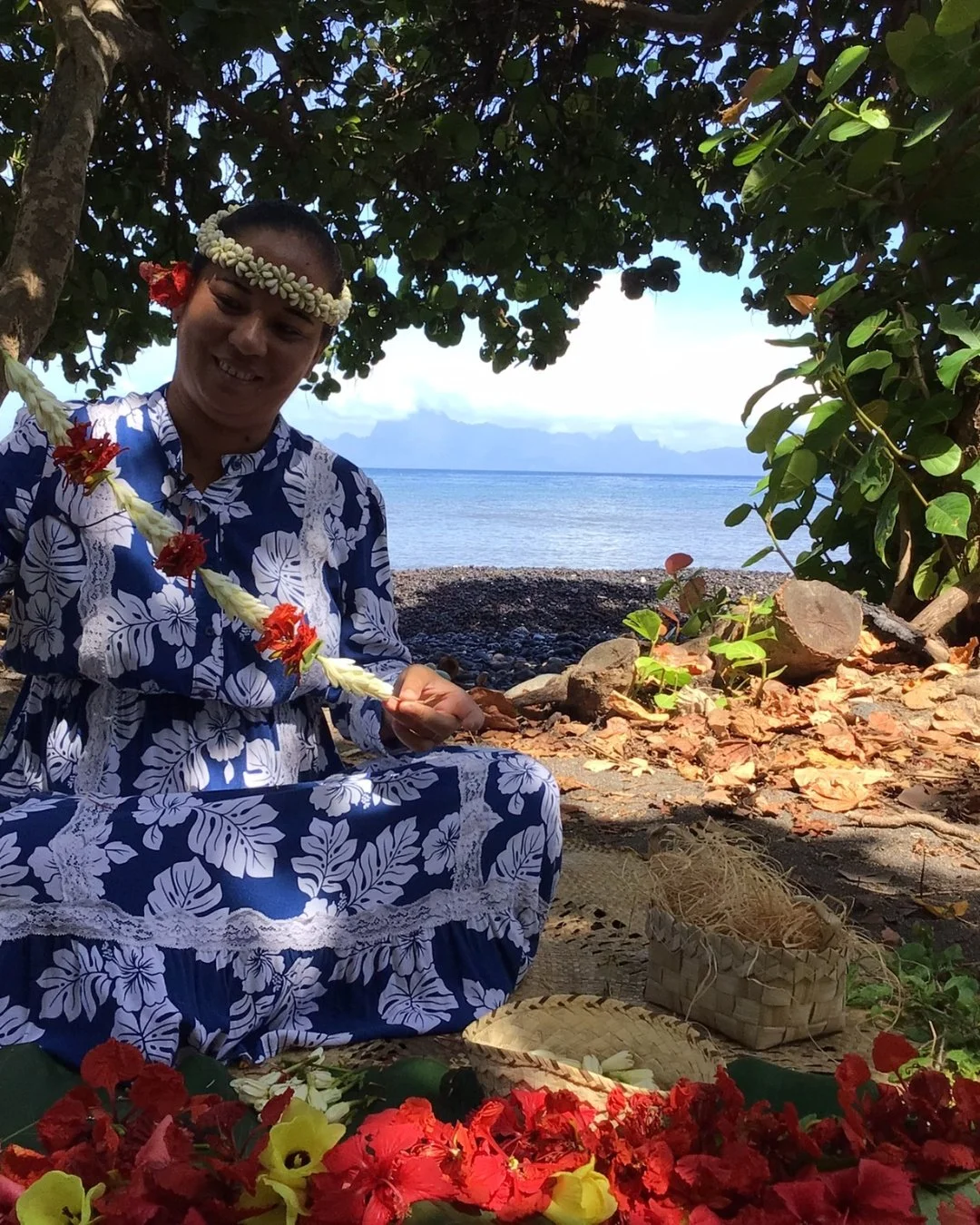 Une femme en tenue traditionnelle pose sur une plage, portant une couronne de fleurs, entourée de fleurs colorées, avec la mer en toile de fond et des arbres ombragés.