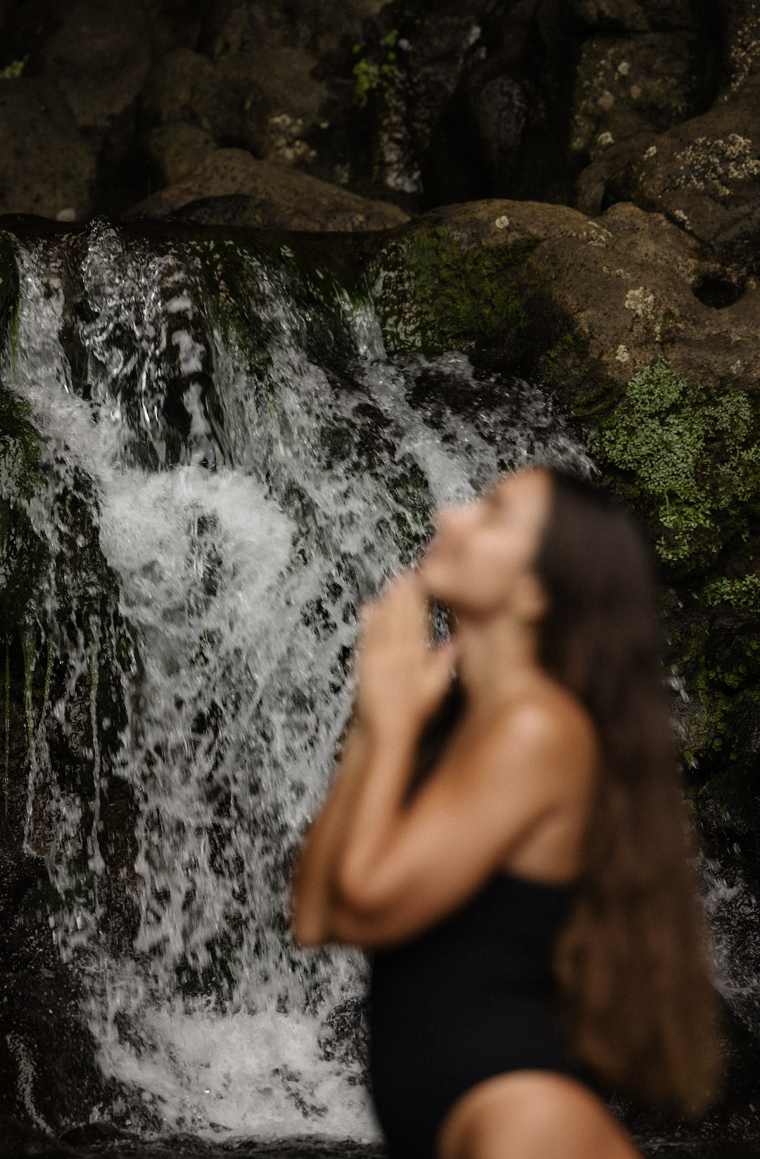Une femme en maillot noir devant une cascade de montagne, floue, avec des rochers et de la végétation en arrière-plan.