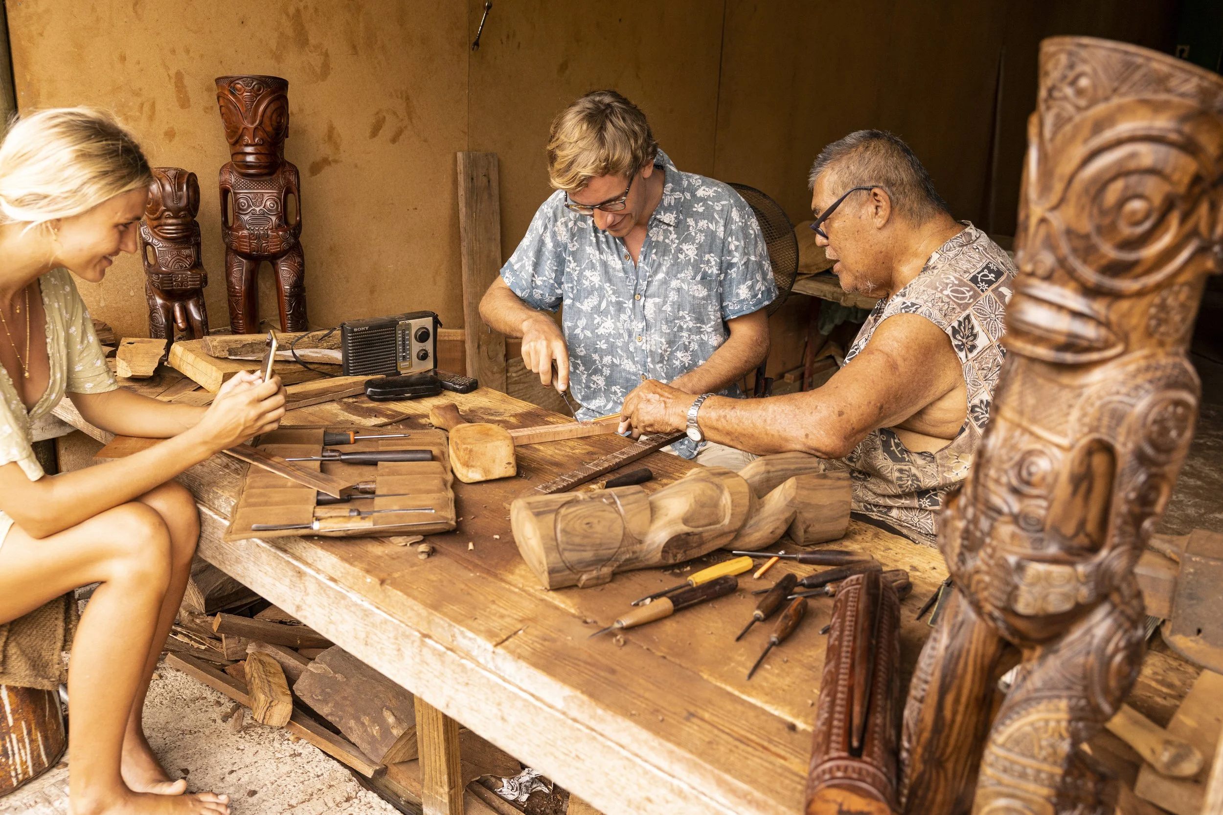 Groupe de trois personnes travaillant sur des sculptures en bois traditionnelles polynésiennes dans un atelier en bois. Autour d'eux, des outils et des statues en bois décoratives.