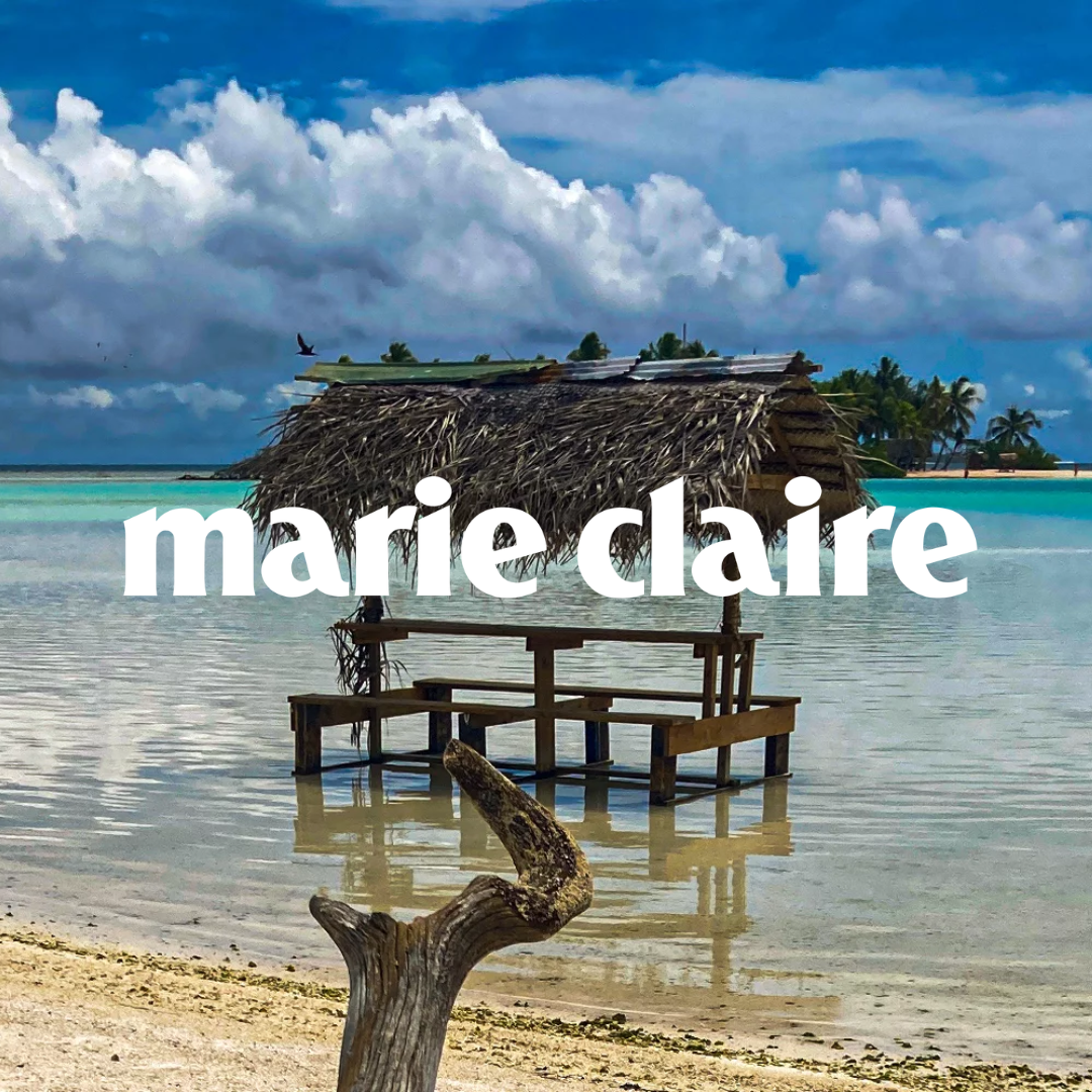 Un cabanon en bois avec un toit en feuilles de palmier sur une plage de sable, avec des eaux turquoise, des palmiers, et un ciel nuageux.