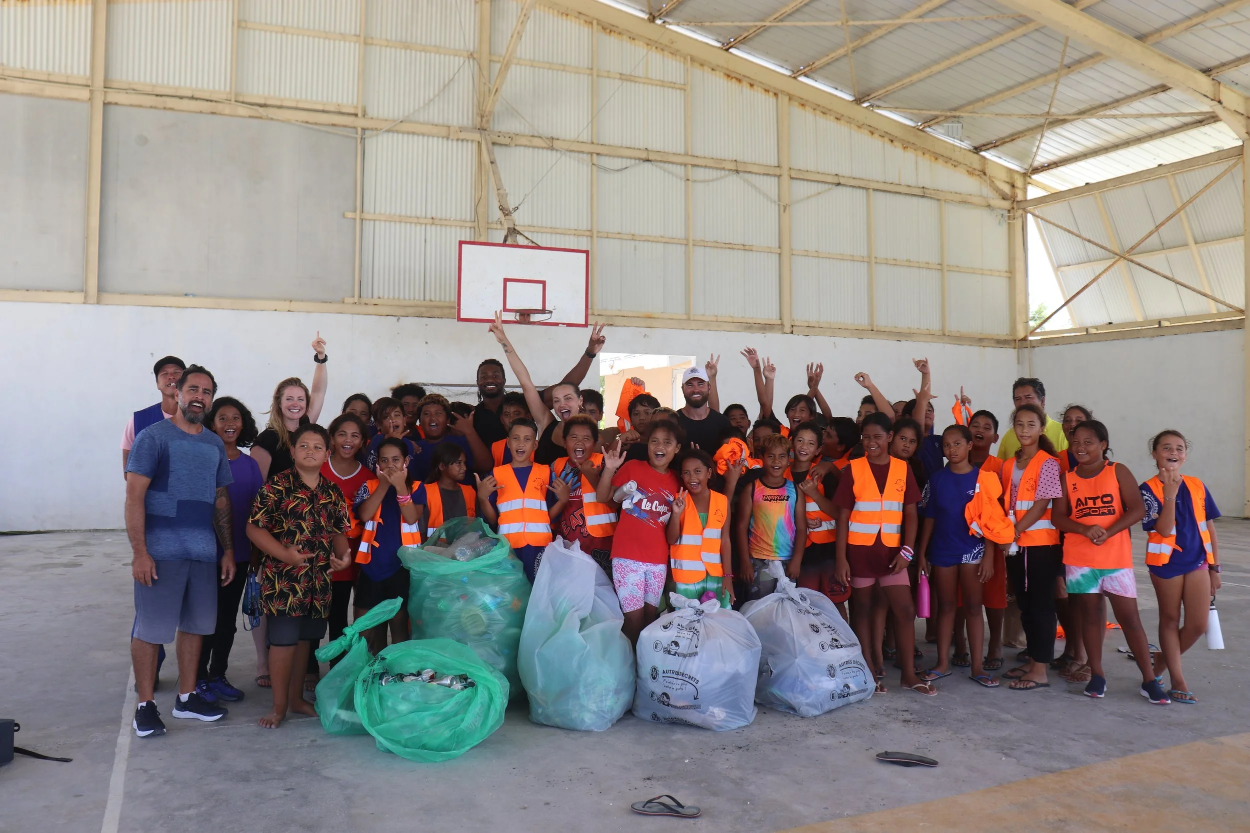 Groupe d'enfants et d'adultes dans un gymnase, posant avec des sacs remplis de déchets après une opération de nettoyage.