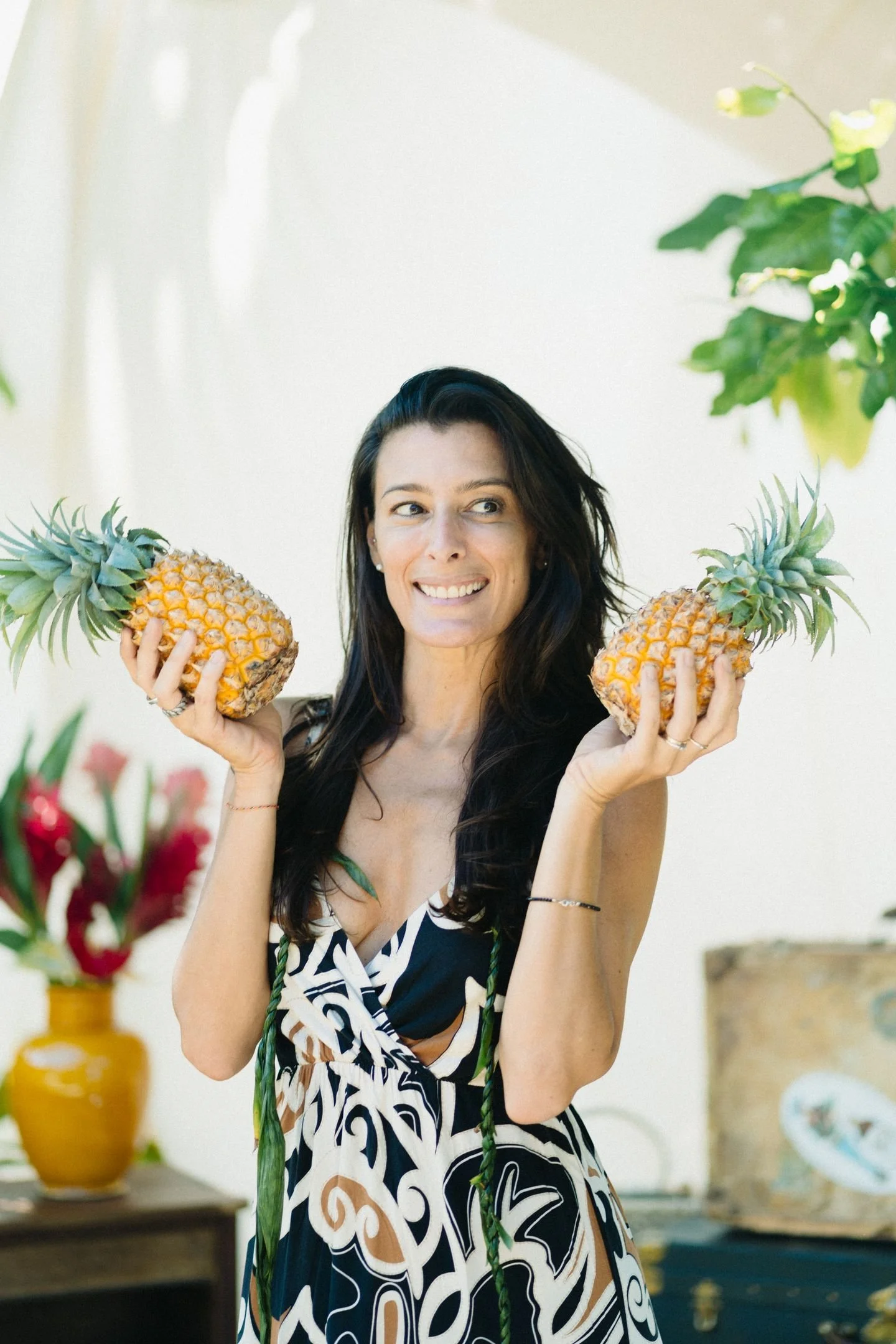Une femme souriante tient deux ananas, entourée de décorations florales et de vases colorés dans un intérieur lumineux.