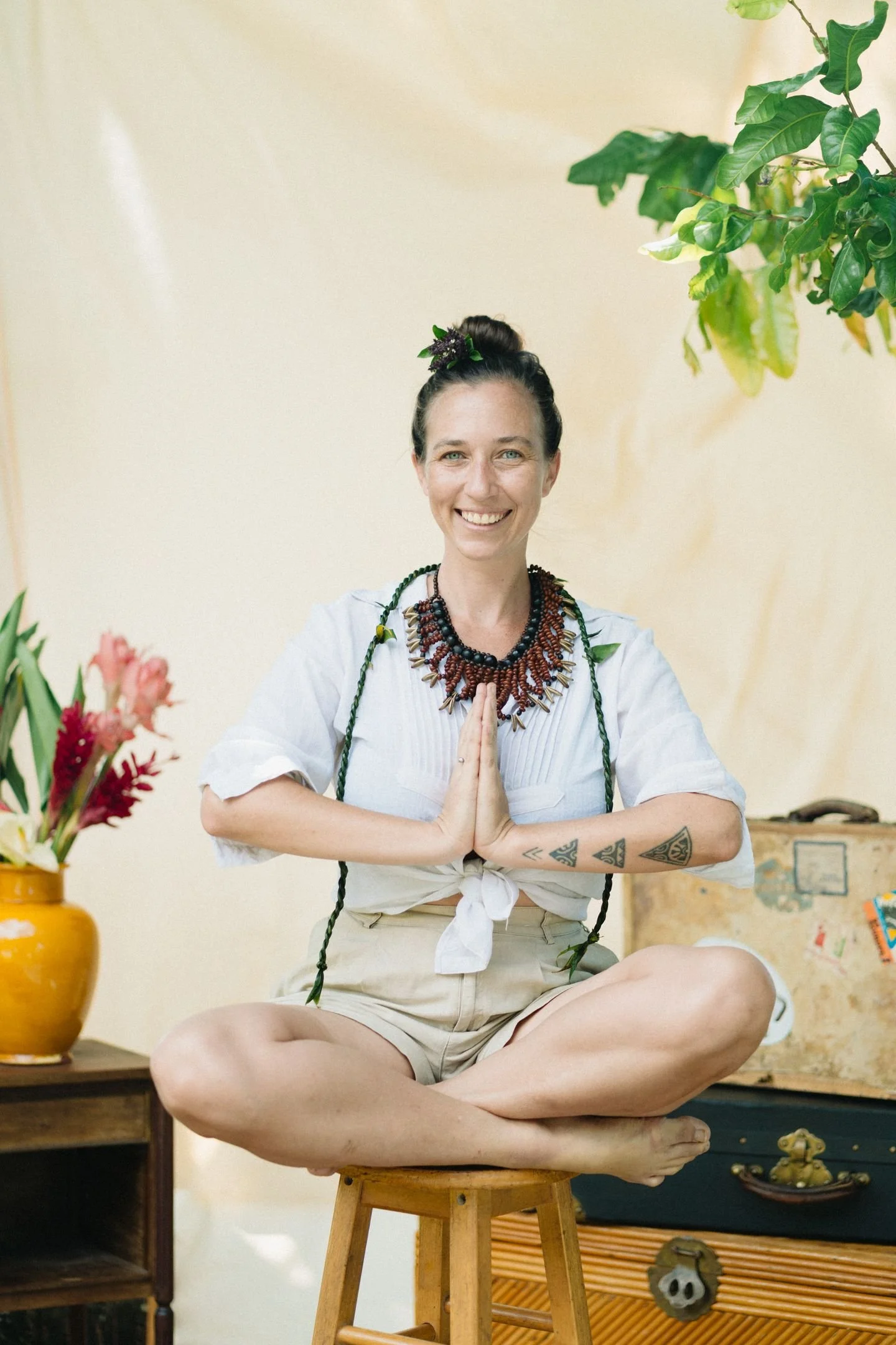 Une femme assise en position de yoga en méditation, faisant un geste de prière avec ses mains, portant des bijoux et des vêtements décontractés, avec un décor de fleurs et de meubles en arrière-plan.