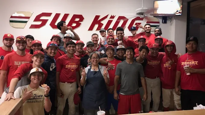 Group of young men and women in red and gray shirts, some making peace signs, standing in front of a Subway Kings sign inside a restaurant.