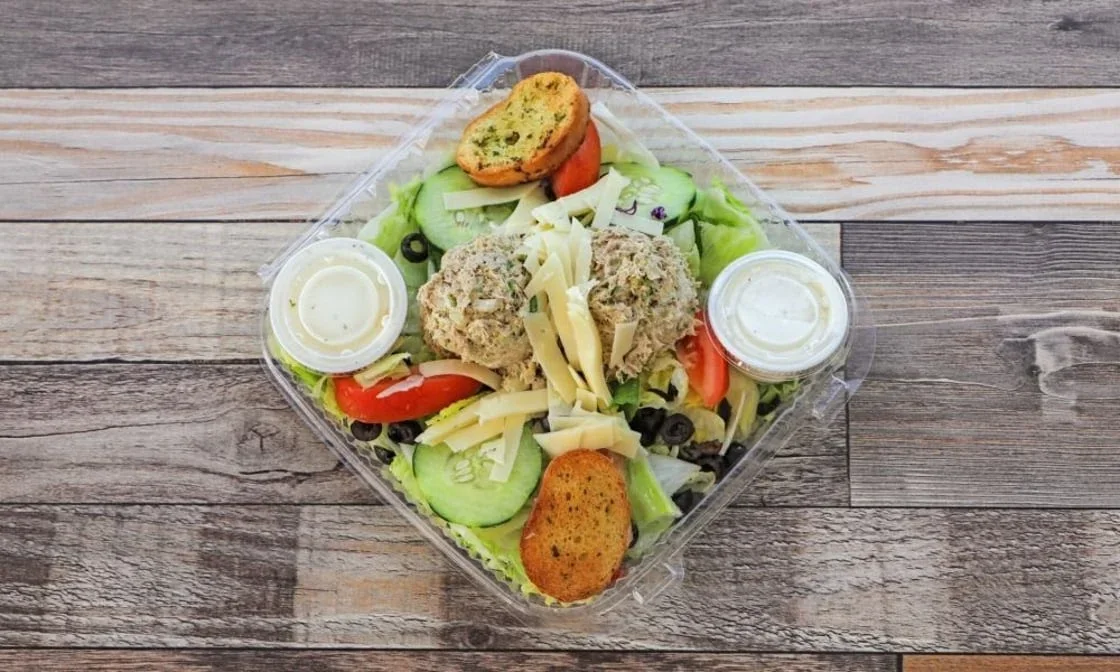 Checkers salad with lettuce, sliced cucumbers, cherry tomatoes, black olives, shredded cheese, two chicken salad scoops, two breaded fried items, two containers of dressing, and a toasted bread slice, all in a clear plastic container on a wooden surf