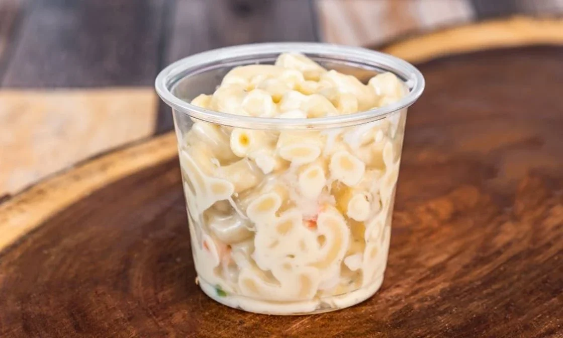 A clear plastic cup filled with macaroni salad containing pasta, mayonnaise, and chopped vegetables, sitting on a wooden surface.