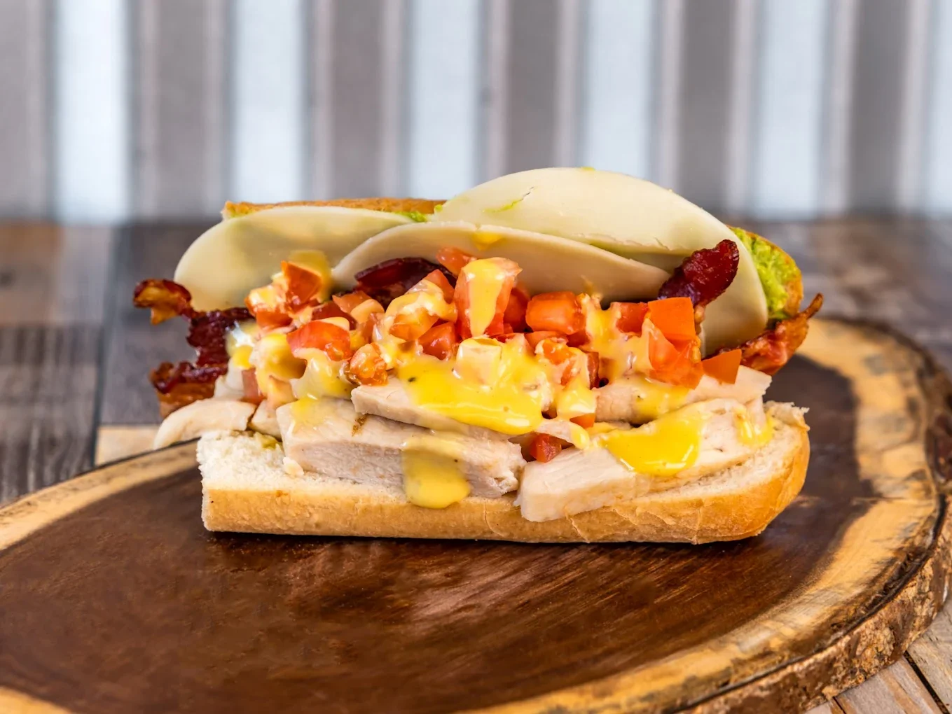 Open-faced sandwich with turkey, bacon, scrambled eggs, cheese, tomato, and pickles on a toasted bun on a wooden plate.