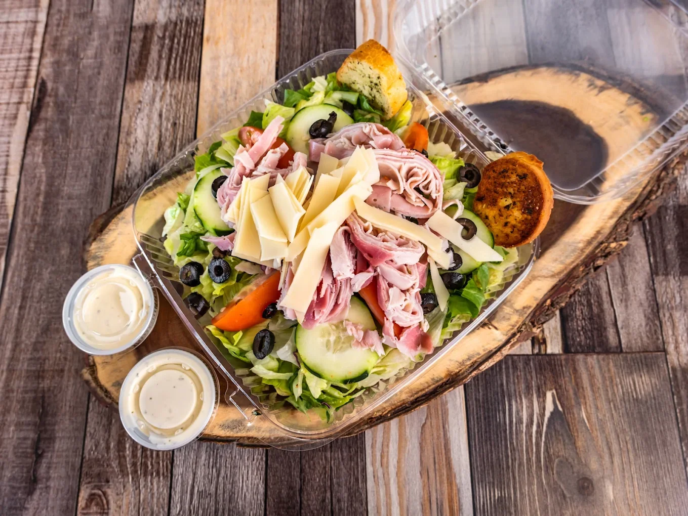 Fresh deli salad with lettuce, cucumbers, tomatoes, black olives, and various cheeses, accompanied by two small cups of dressing and two bread rolls, on a rustic wooden board.