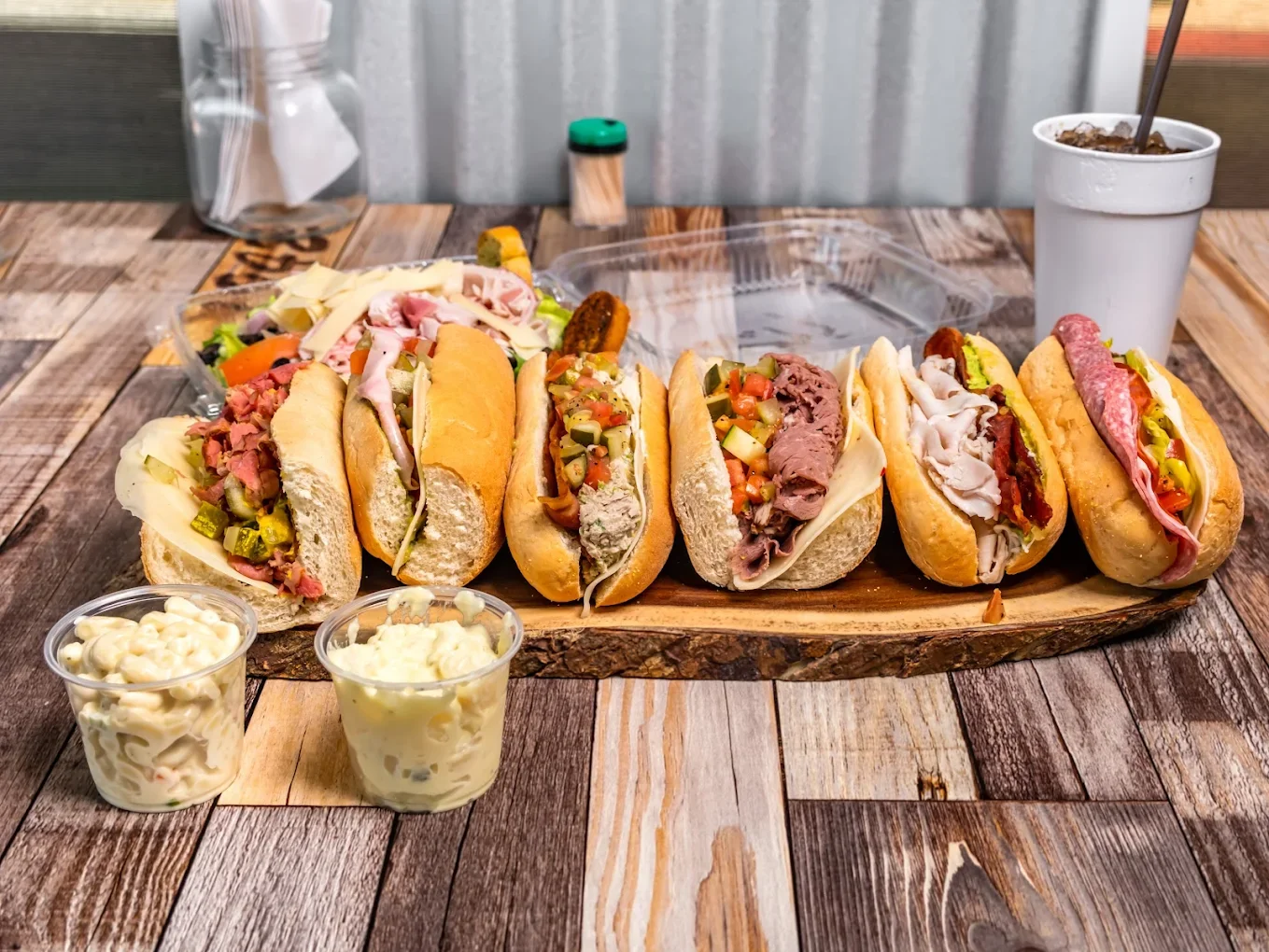 A variety of seven hot dogs in buns with different toppings, served on a wooden platter, with two cups of macaroni and cheese, and a cup of soda with ice, on a rustic wooden table.