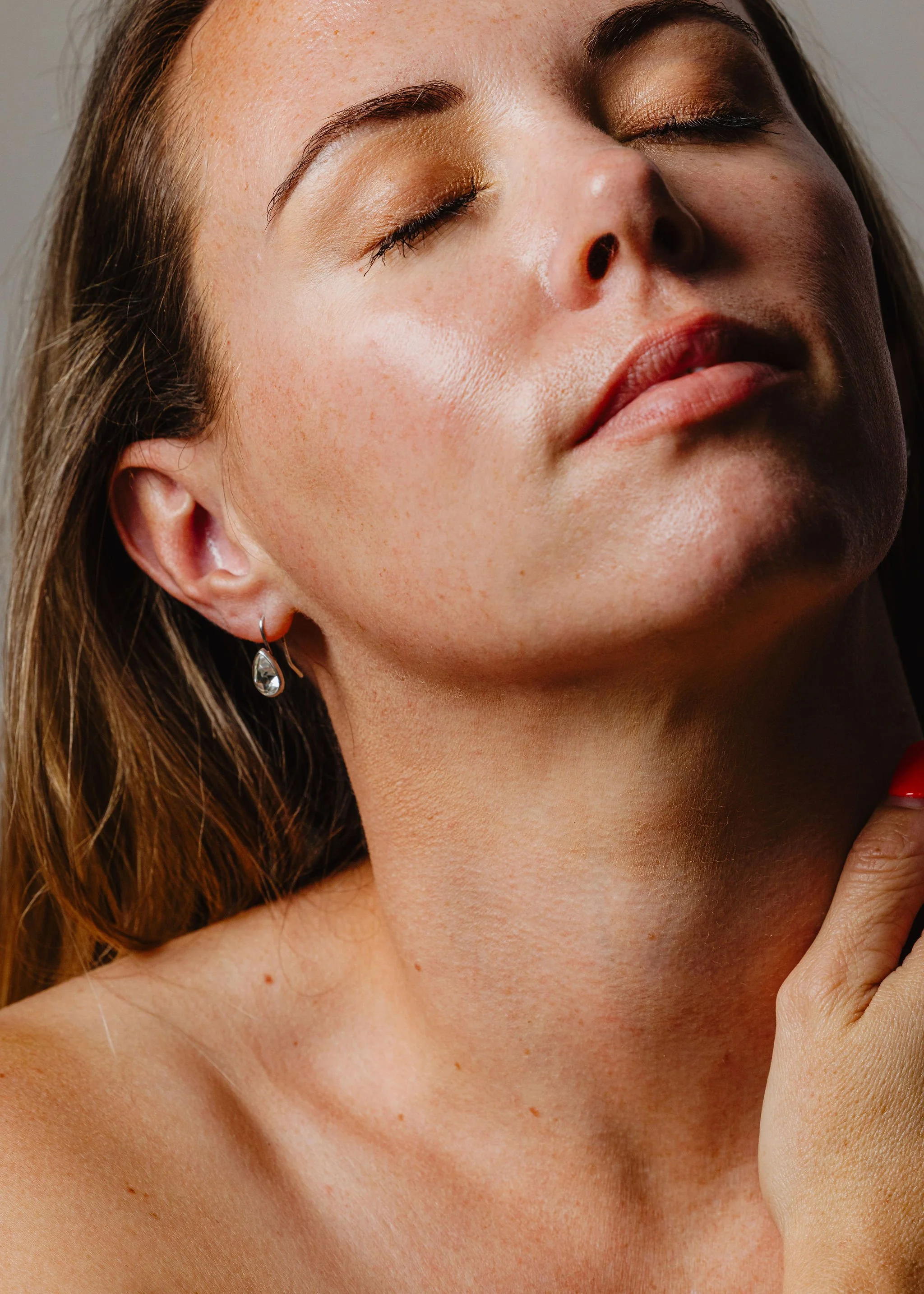 Close-up of a woman with closed eyes, wearing gold eyeshadow and earrings, touching her neck gently.