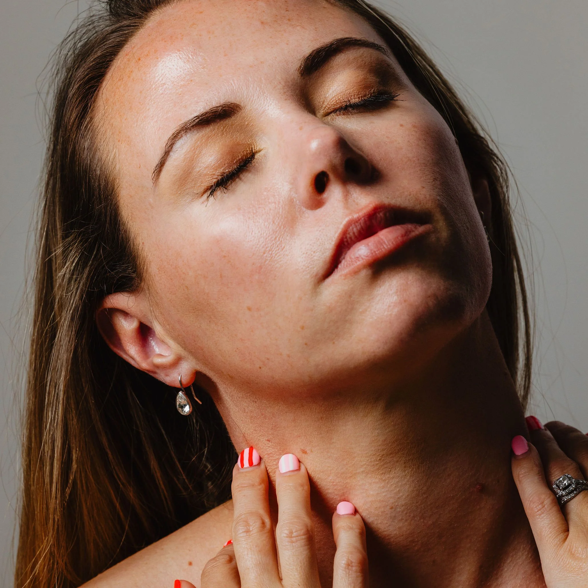 Close-up of a woman with fair skin and freckles, eyes closed, touching her shoulder, featuring multicolored striped and pink nail polish, wearing earrings, and with a watch on her wrist.