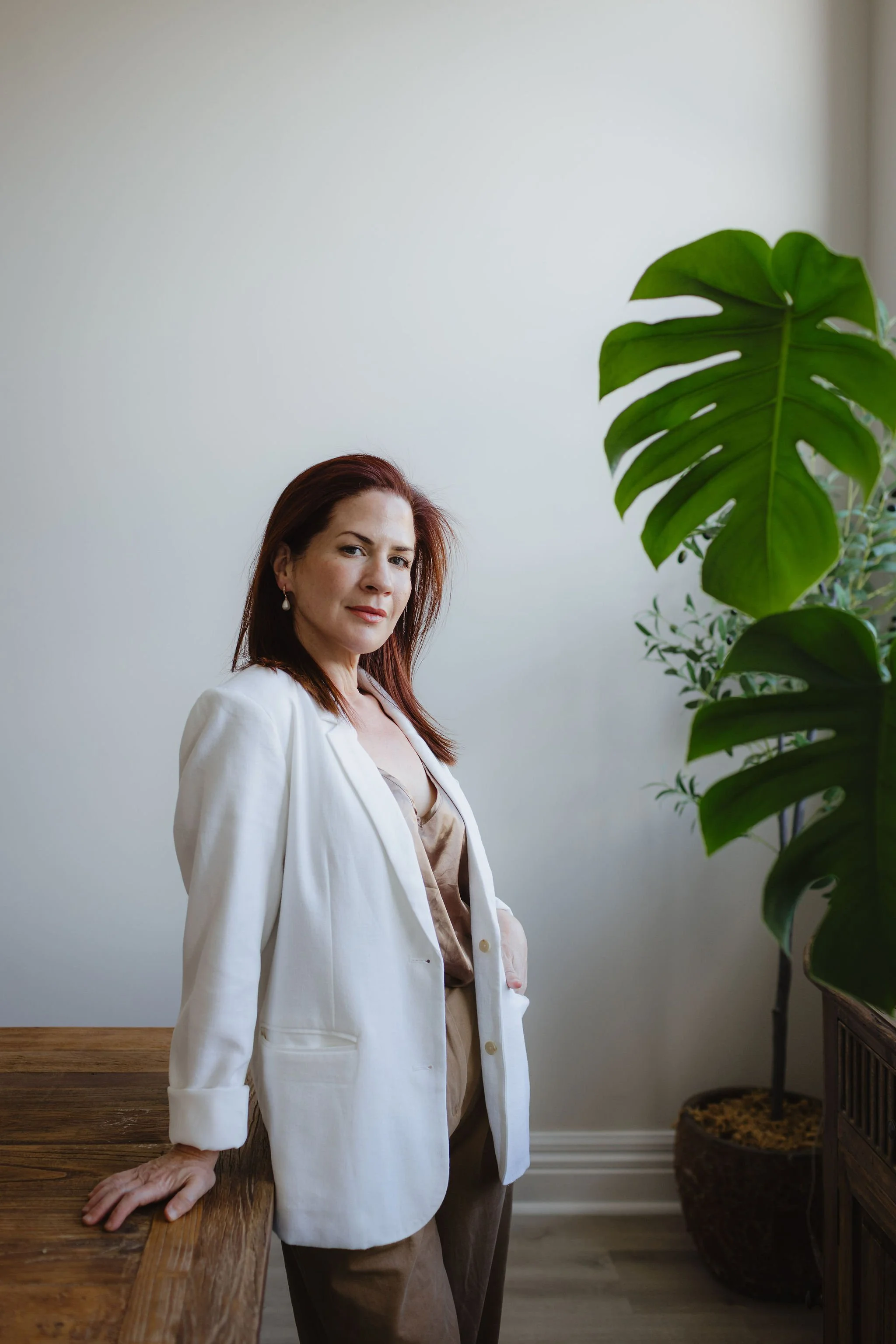 A woman with auburn hair and earrings, wearing a white blazer and beige top, leaning on a wooden table with her hand, standing in a room with a large potted plant with broad green leaves.