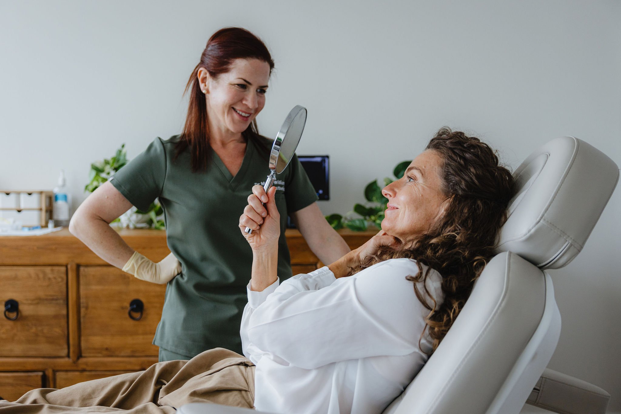 A nurse in green scrubs holding a mirror to a smiling woman lying in a medical chair, with a wooden cabinet and plants in the background.