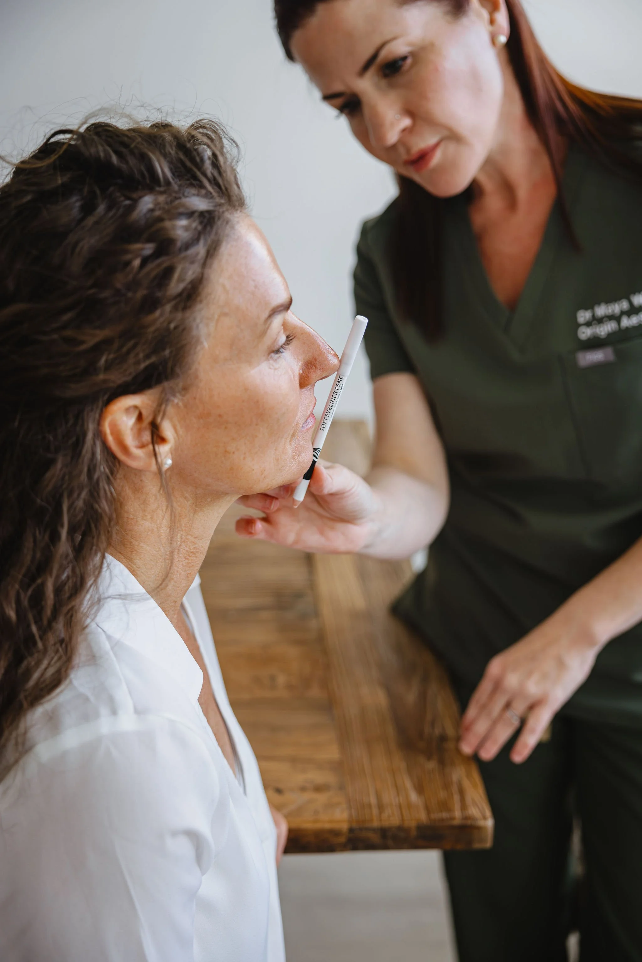 A woman with long brown hair and a white uniform appears to be preparing for a medical examination or procedure in a clinical setting. She is holding a soft eyeliner pencil near a patient’s face, who is lying on a wooden table. The staff member has a focused expression, and her uniform has a name tag that partially reads "Dr. Mary...".