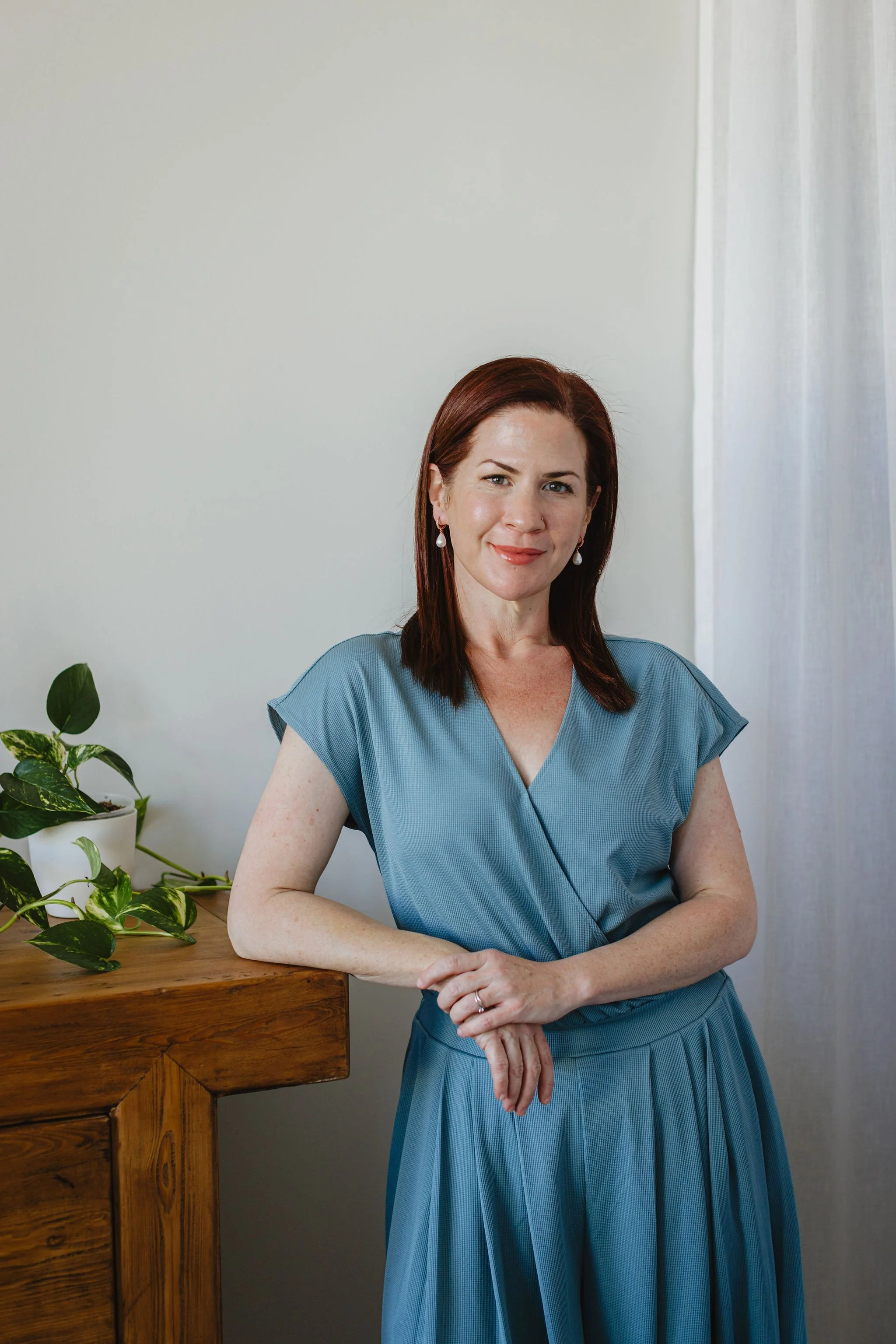 A woman with shoulder-length reddish-brown hair, wearing a blue dress and pearl earrings, sitting with her hands clasped on a wooden table next to a potted plant, smiling at the camera against a plain light-colored wall.