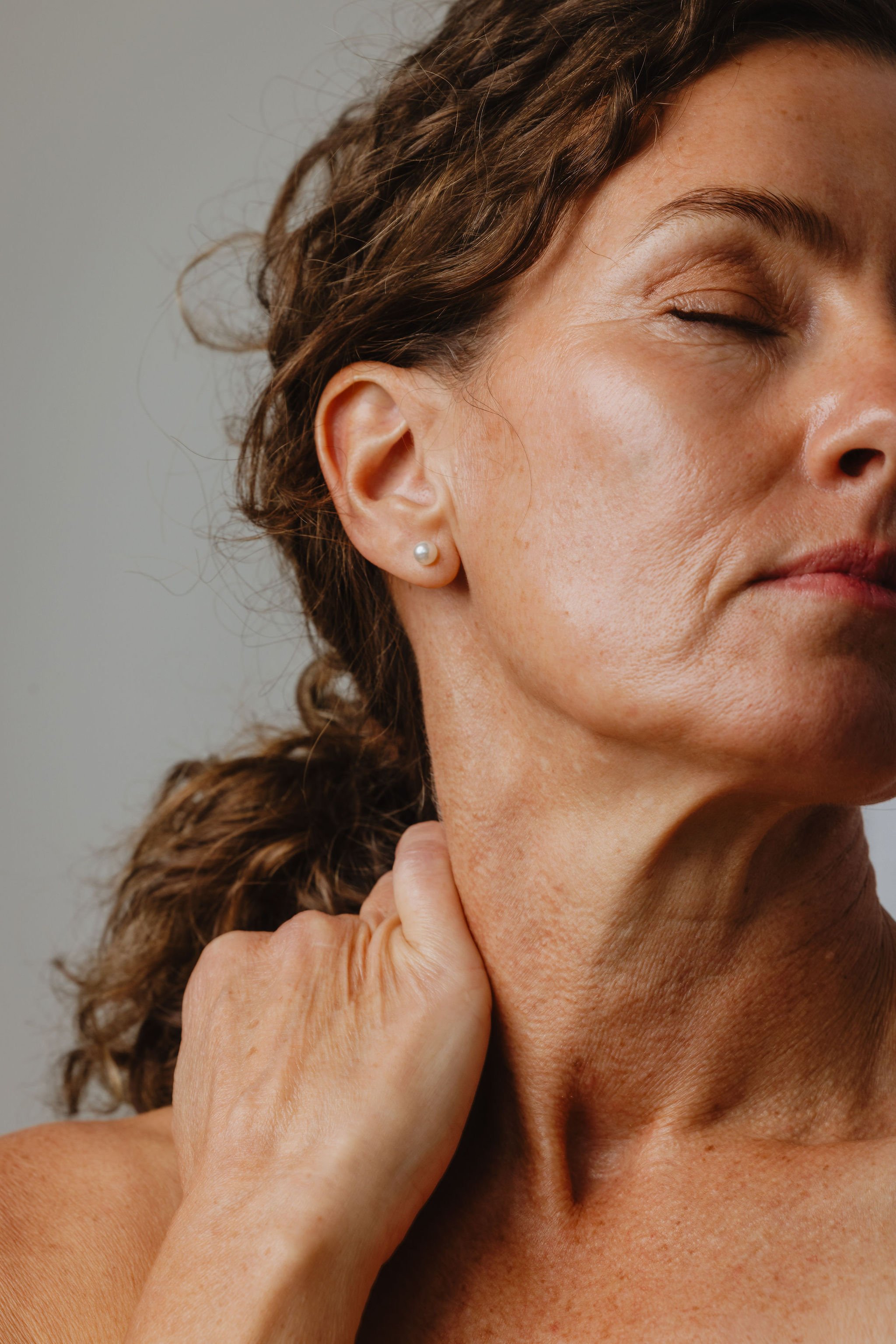 Close-up of a woman with closed eyes, brown curly hair, wearing a pearl earring, touching her neck with her hand, showing smooth skin and natural complexion.