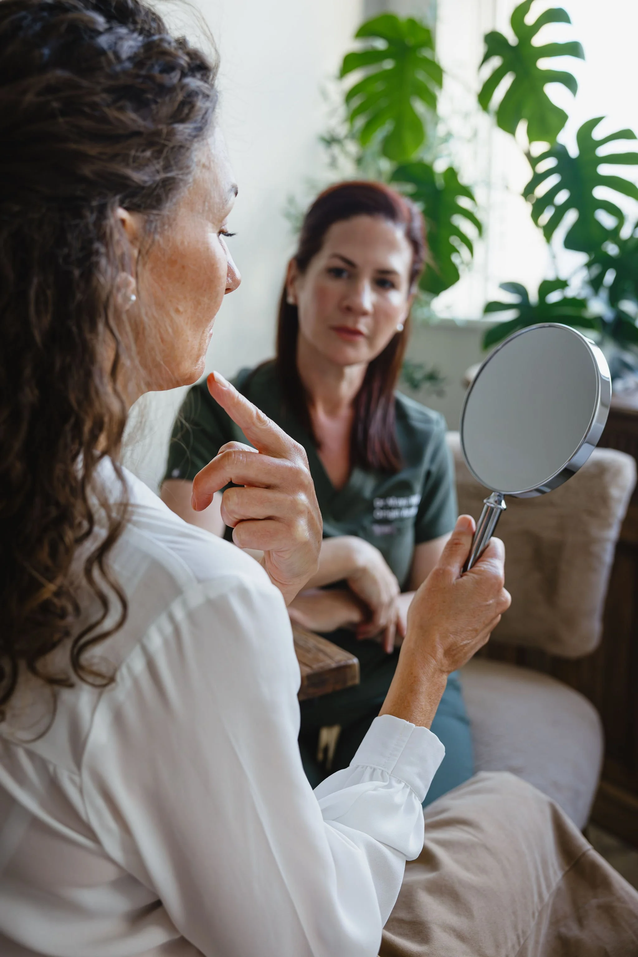 A woman in a white shirt pointing to her face while looking into a hand mirror held by another woman with brown hair, in a room with large green houseplants in the background.