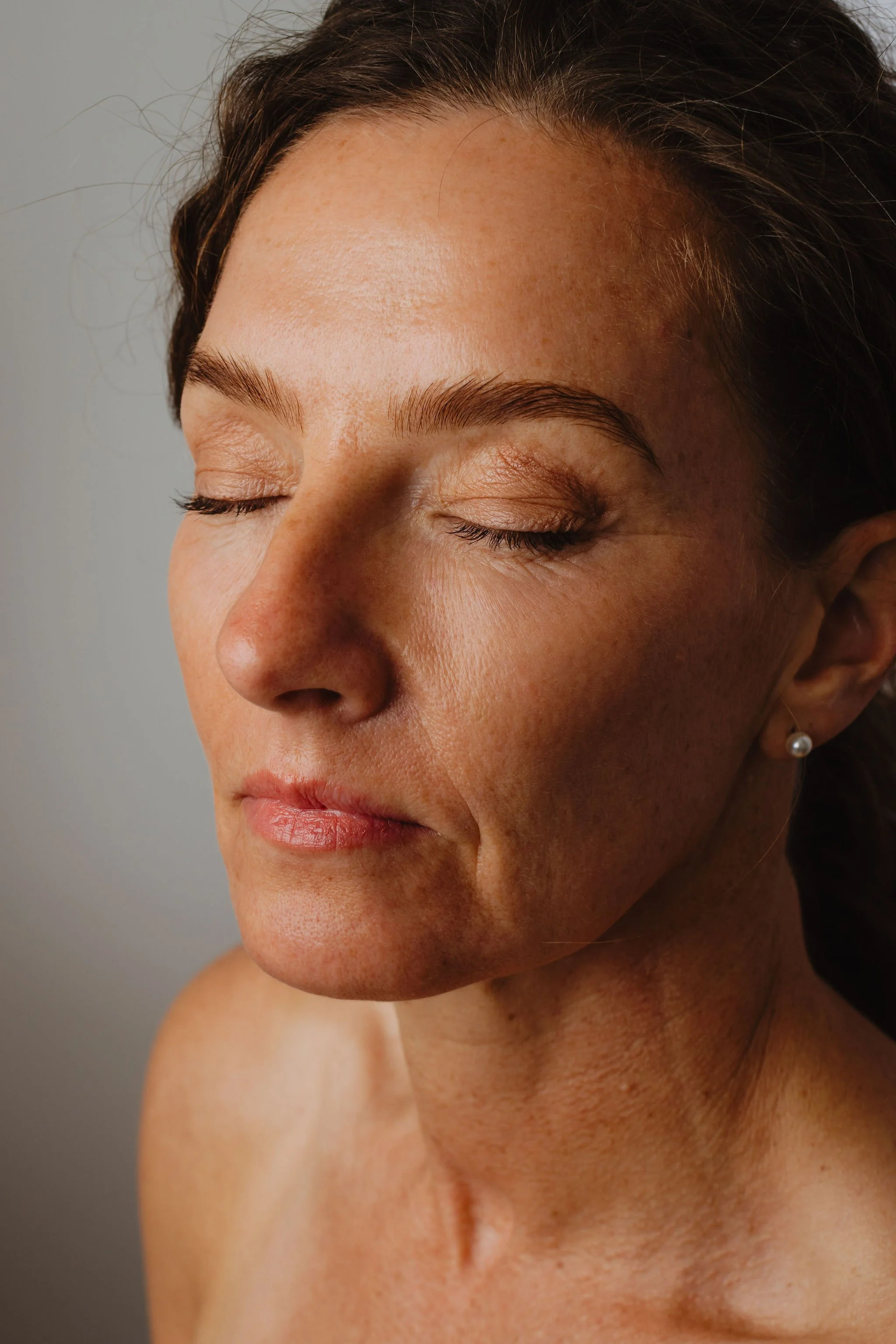 Close-up of a woman's face with closed eyes, showing natural skin texture and dark hair, wearing a pearl earring.