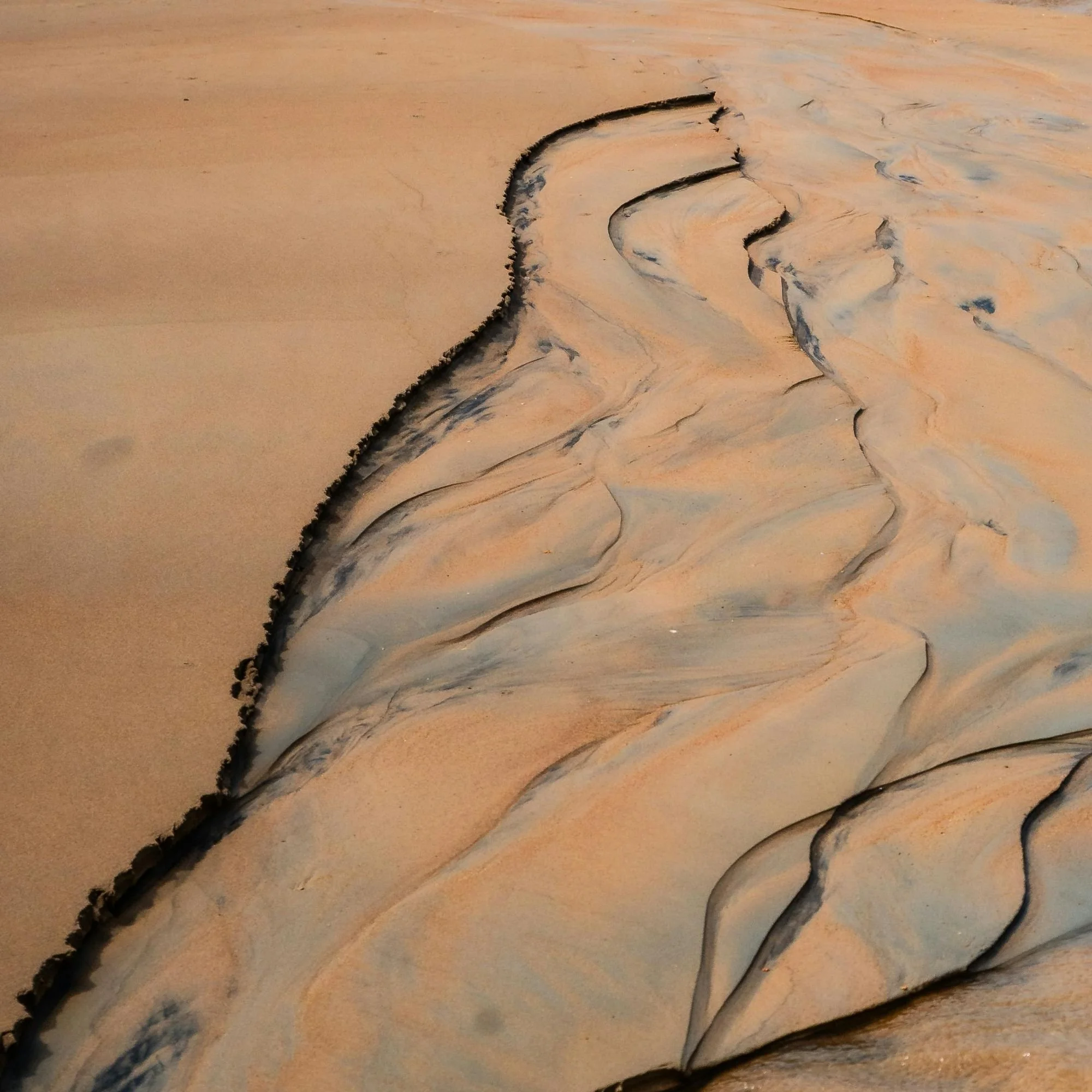 Close-up photograph of sand dunes with rippled patterns and a clear sky. The dunes have a mix of light and dark sandy shades.