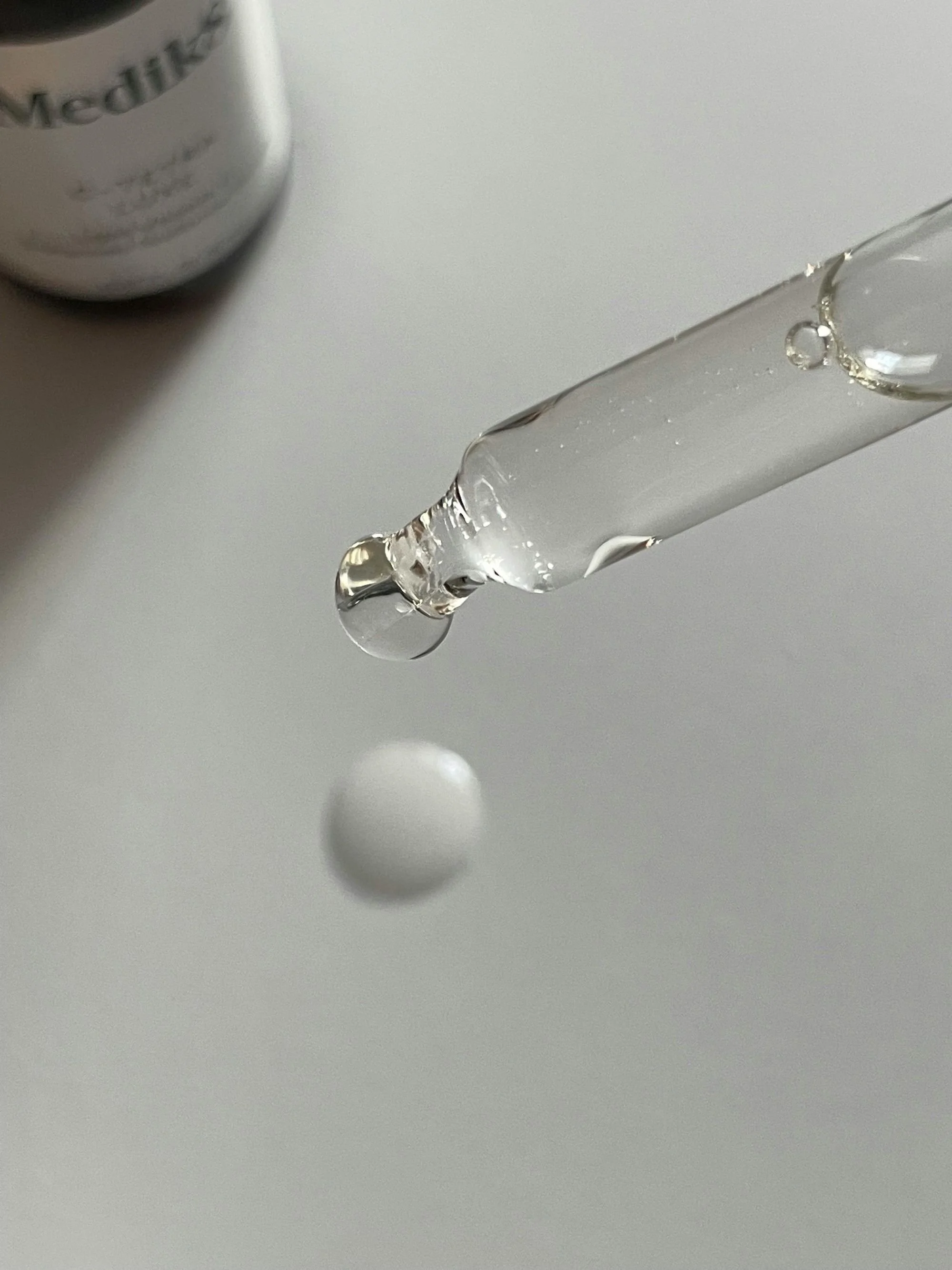 Close-up of a dropper releasing a droplet of clear liquid with a blurred medication bottle in the background.