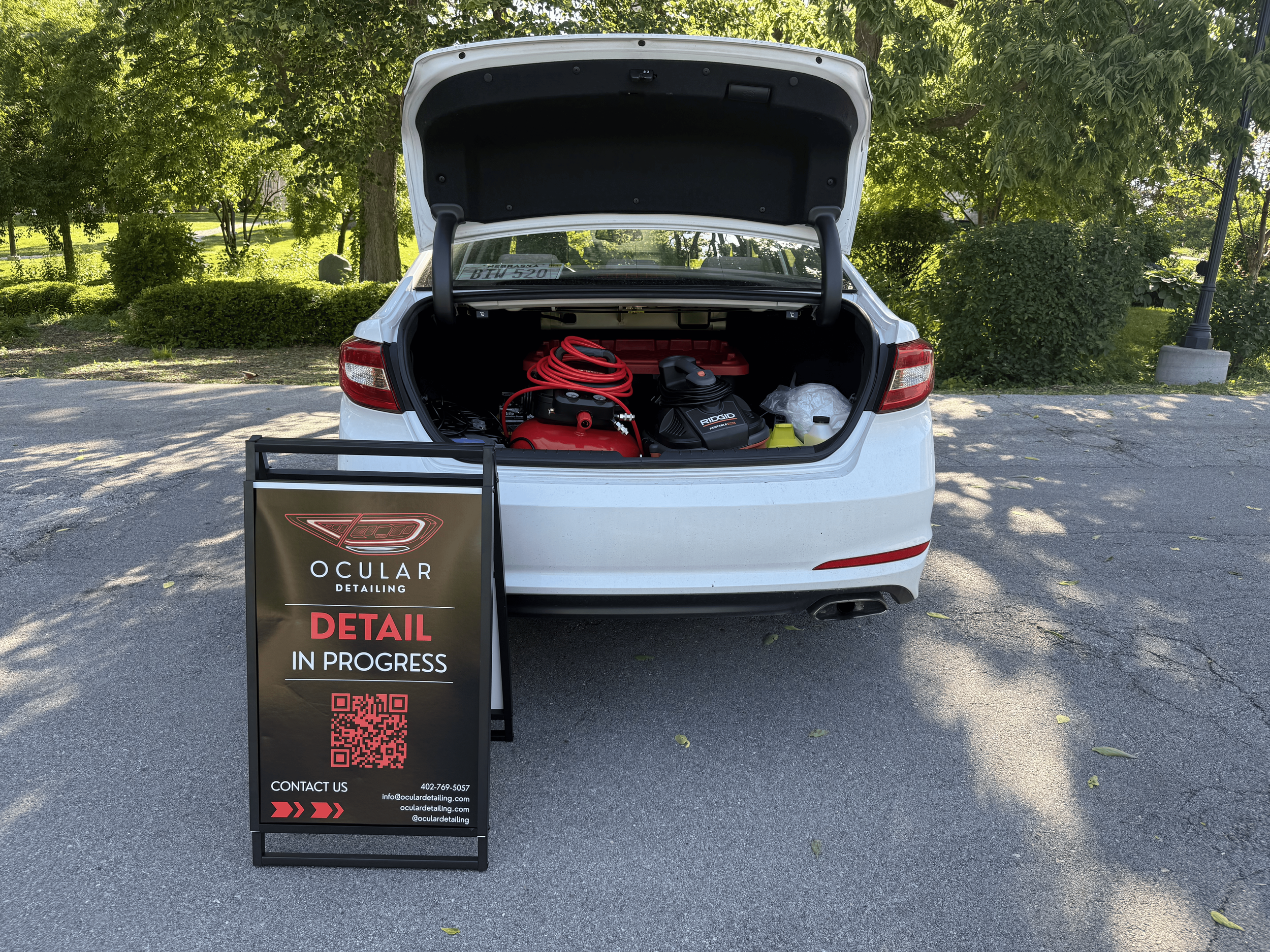 Open trunk of a white car displaying cleaning or detailing equipment including a red and black vacuum, a red extension cord, and a black spray bottle, with a sign in front that reads 'Ocular Detailing: Detail in Progress' and includes contact information and a QR code.