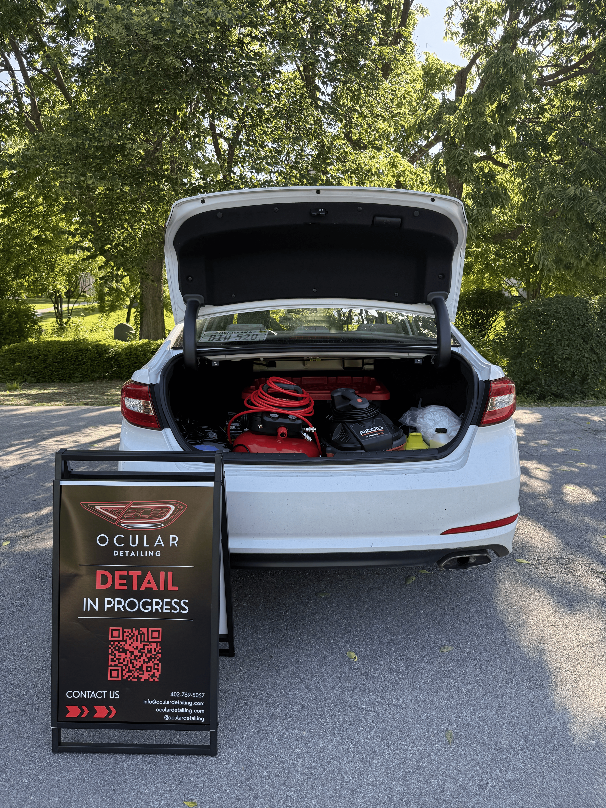 A white car with its trunk open, parked outdoors near green trees and bushes. Inside the trunk, there are equipment including a red air hose and a black cleaning device. In front of the car, there's a black standing sign with information about OCULAR