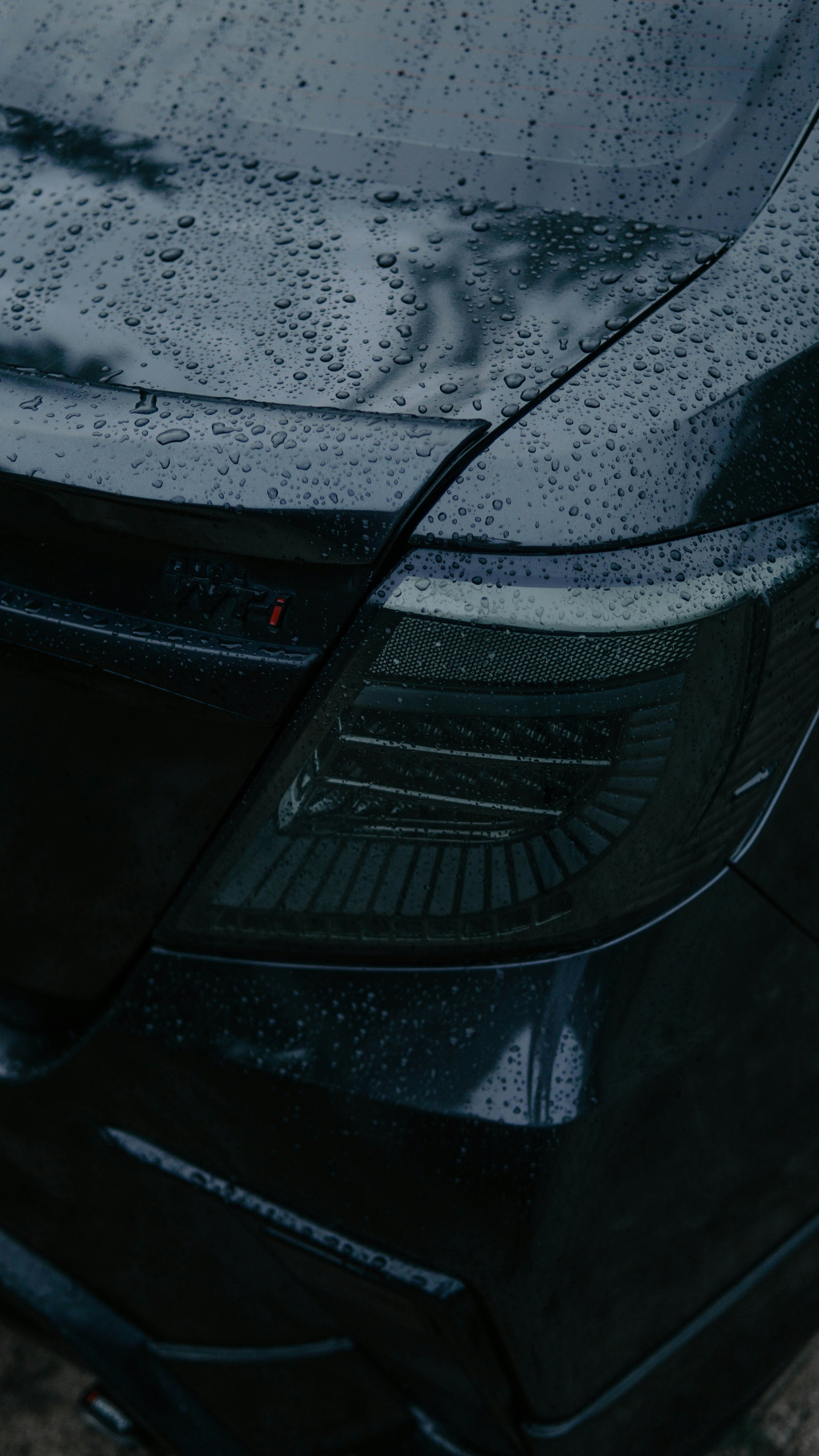 Close-up of a black car's front headlight and hood with raindrops on the surface.
