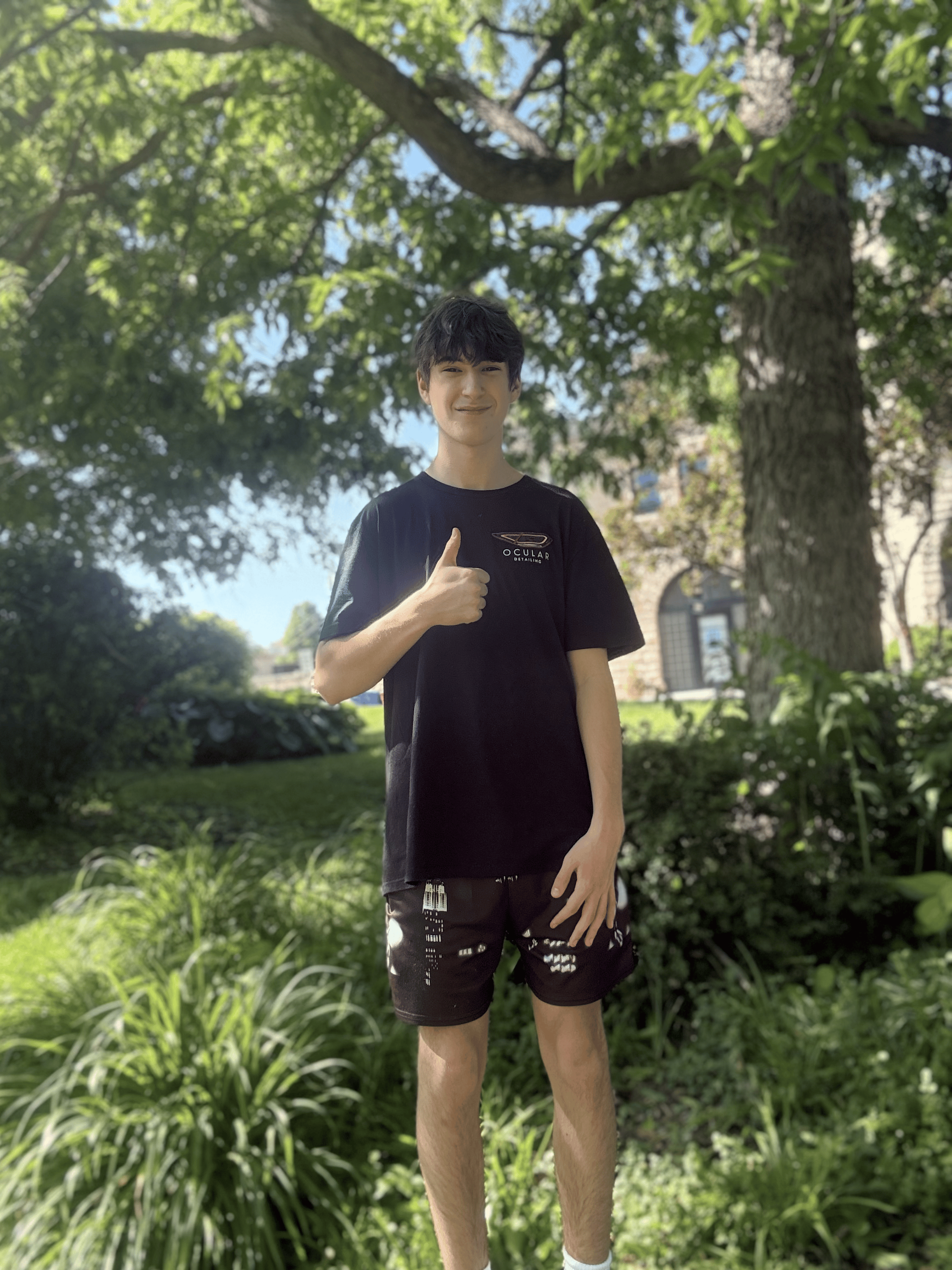 A young man with dark hair smiling and giving a thumbs-up, standing outdoors under a large tree with green leaves, wearing a black T-shirt and black shorts.