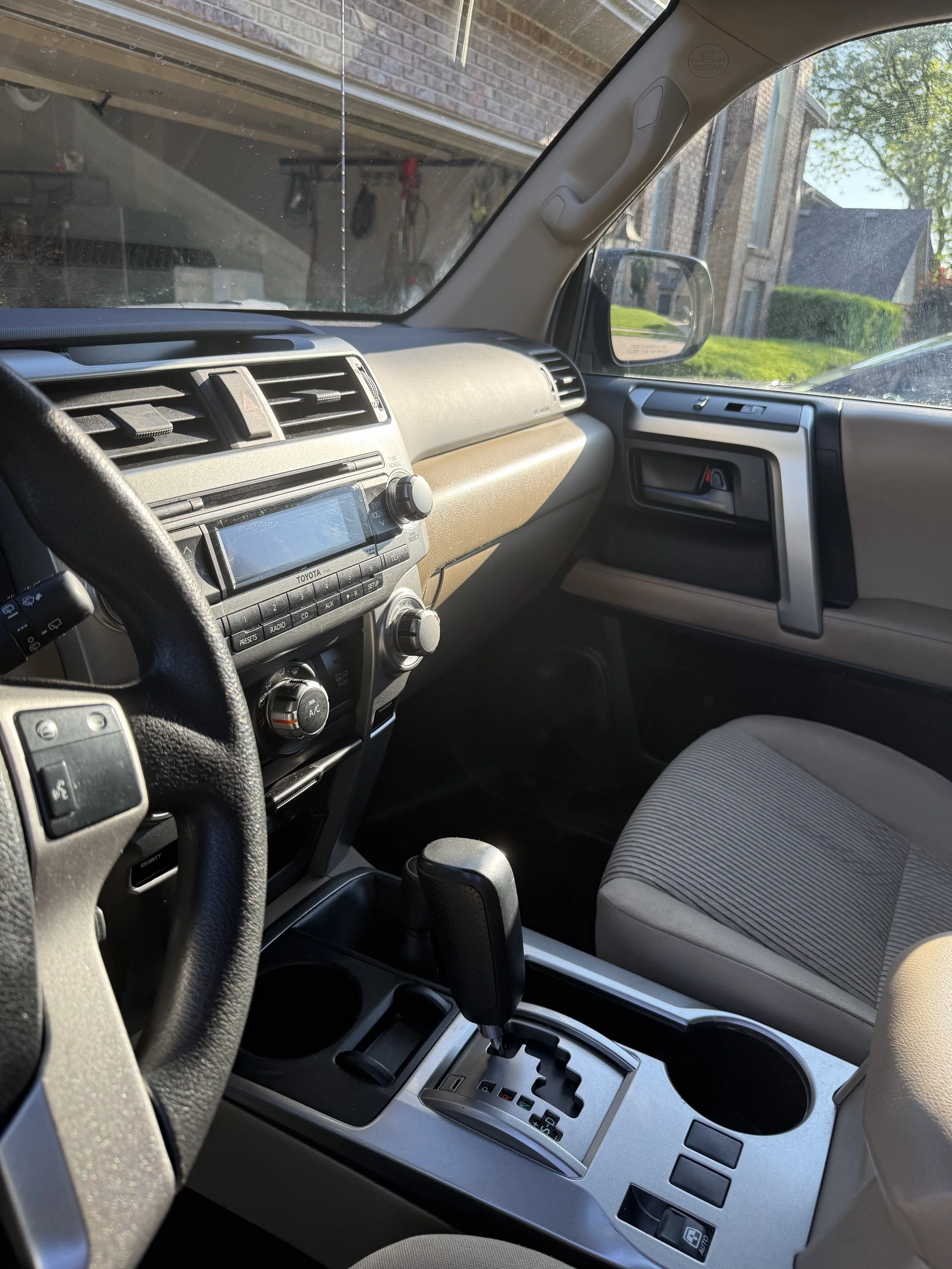 The interior of a car showing the dashboard, steering wheel, center console with gear shift, and front passenger seat. Outside the window, there is a view of a brick house, a garage door, and greenery.