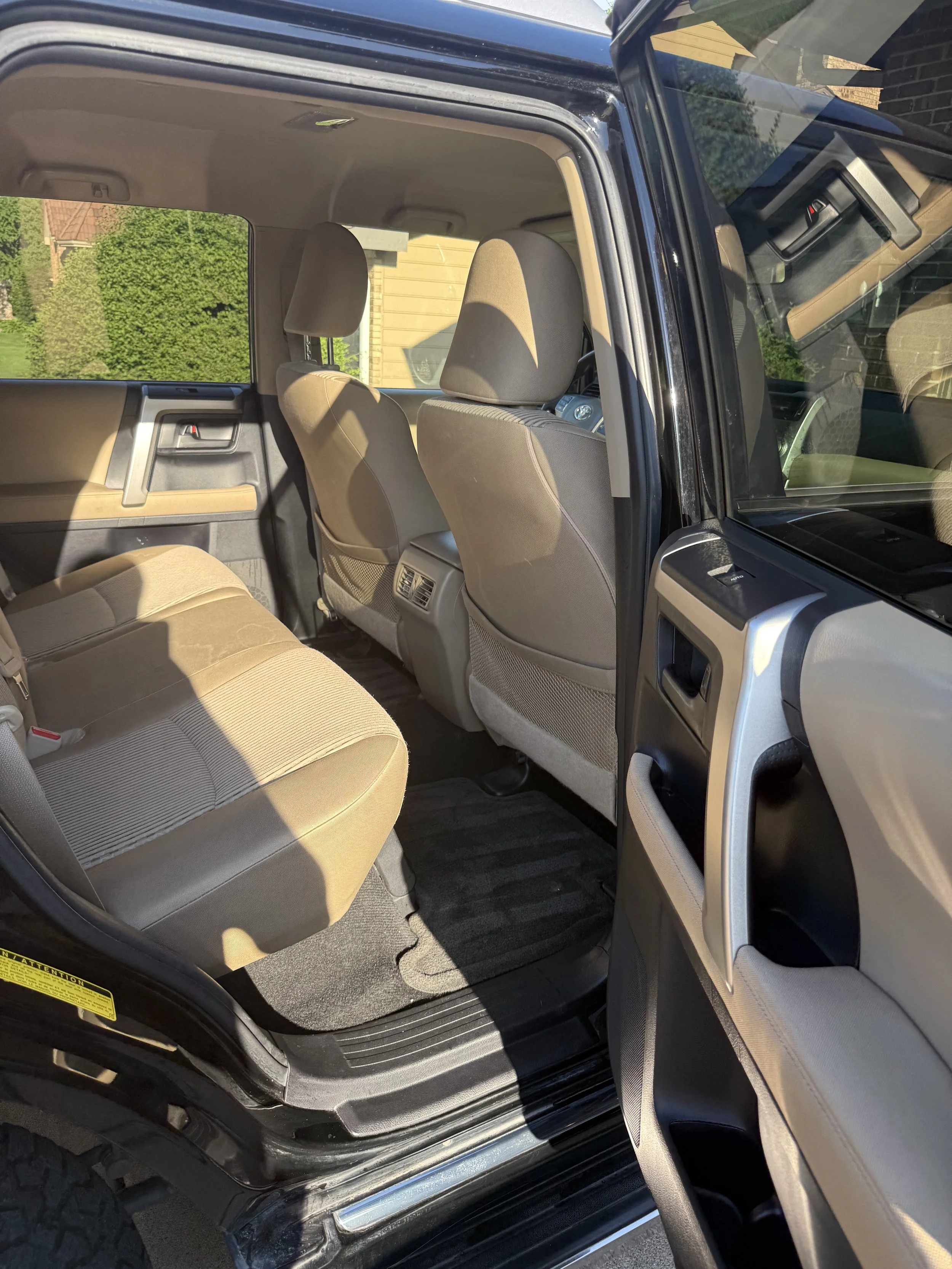 The interior of a vehicle showing beige seats, the back of the front seats, and the dashboard through the open driver's side door. The background includes greenery and a house.