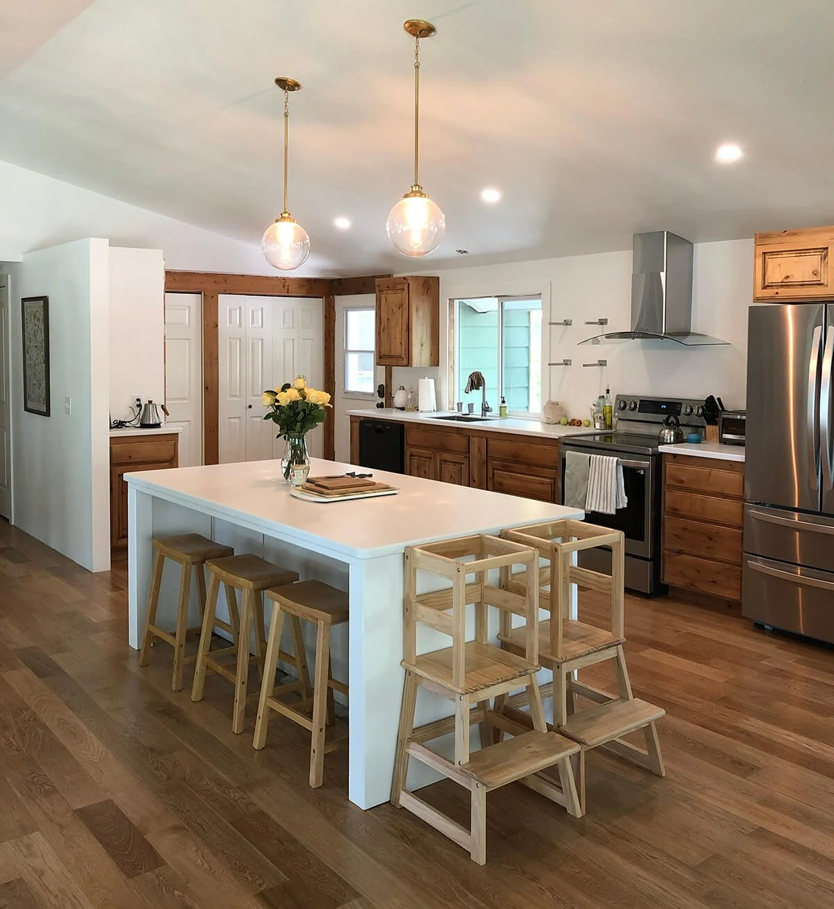 A modern kitchen with wooden cabinets, stainless steel appliances, and a white island. The island has three wooden barstools and a vase with yellow roses. There are two pendant lights hanging from the ceiling, and the floor is hardwood.