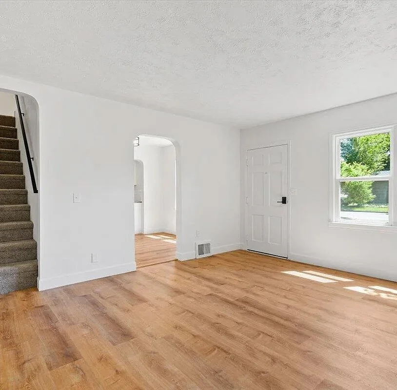 Empty living room with white walls, hardwood floors, a window with green trees outside, a door, and stairs leading upstairs.