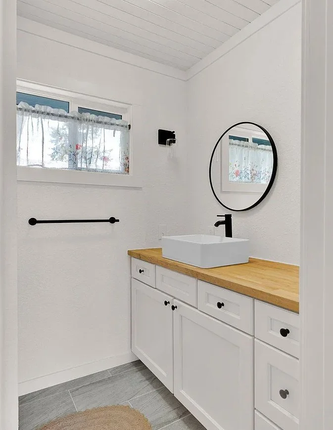 A small bathroom with white walls, a wooden countertop, and a white vessel sink. There's a round mirror above the sink, a small window with a sheer curtain, a black towel bar, and a black wall-mounted light fixture.
