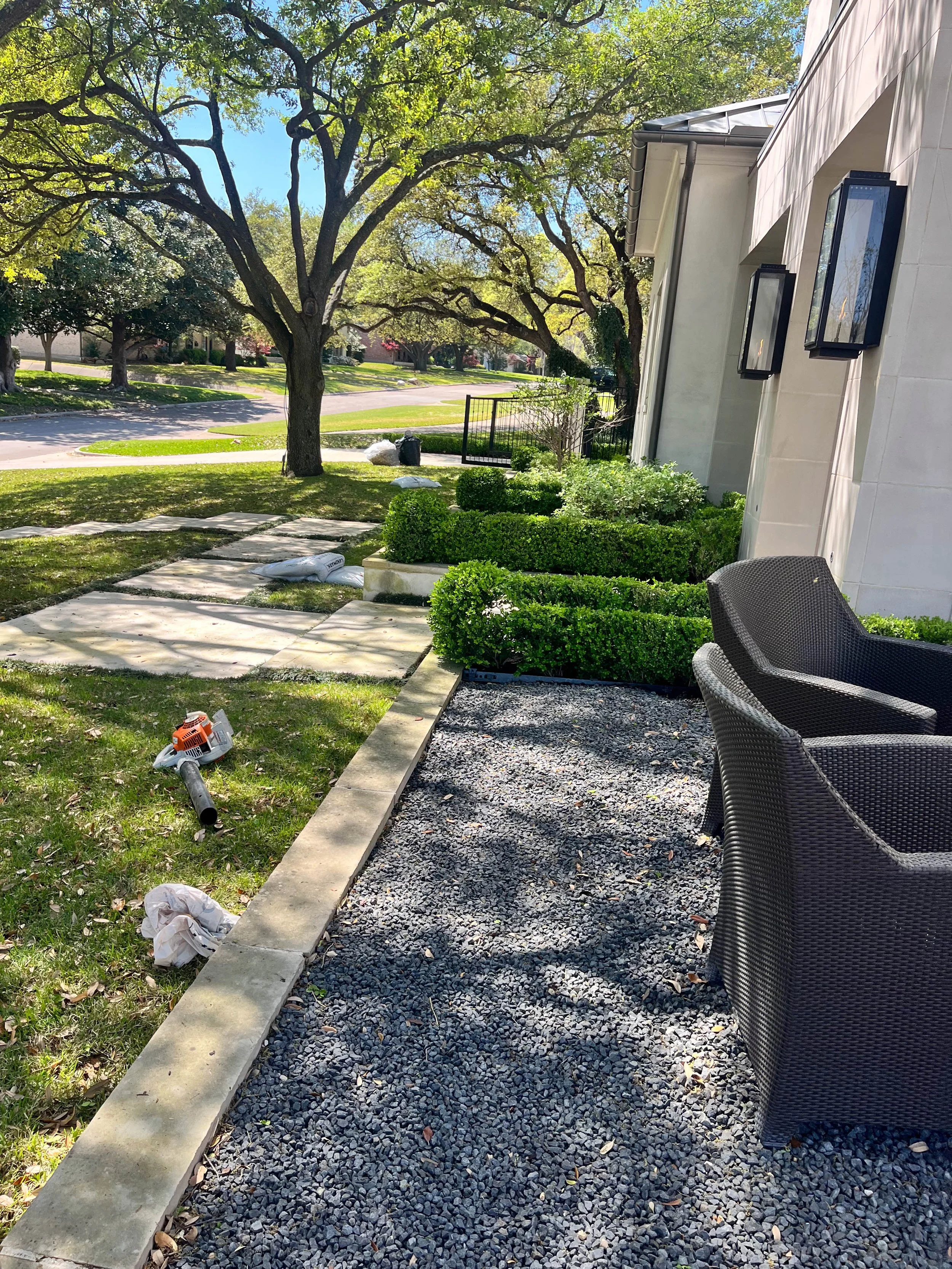 Romero Tree Service project - A backyard patio with a stone pathway, outdoor chairs, green bushes, trees, and a house wall with light-colored siding. There are some bags and a leaf blower on the grass, suggesting yard work.