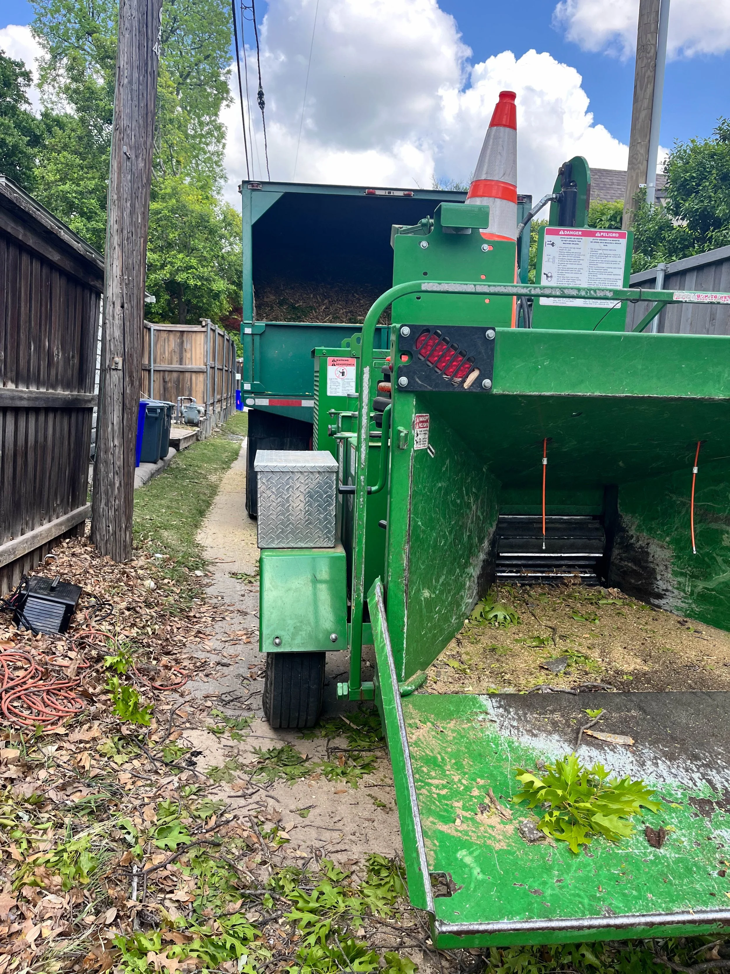 Romero Tree Service green construction cart with orange and white traffic cone on top, parked in a residential backyard with a wooden fence and trees, under a partly cloudy blue sky.