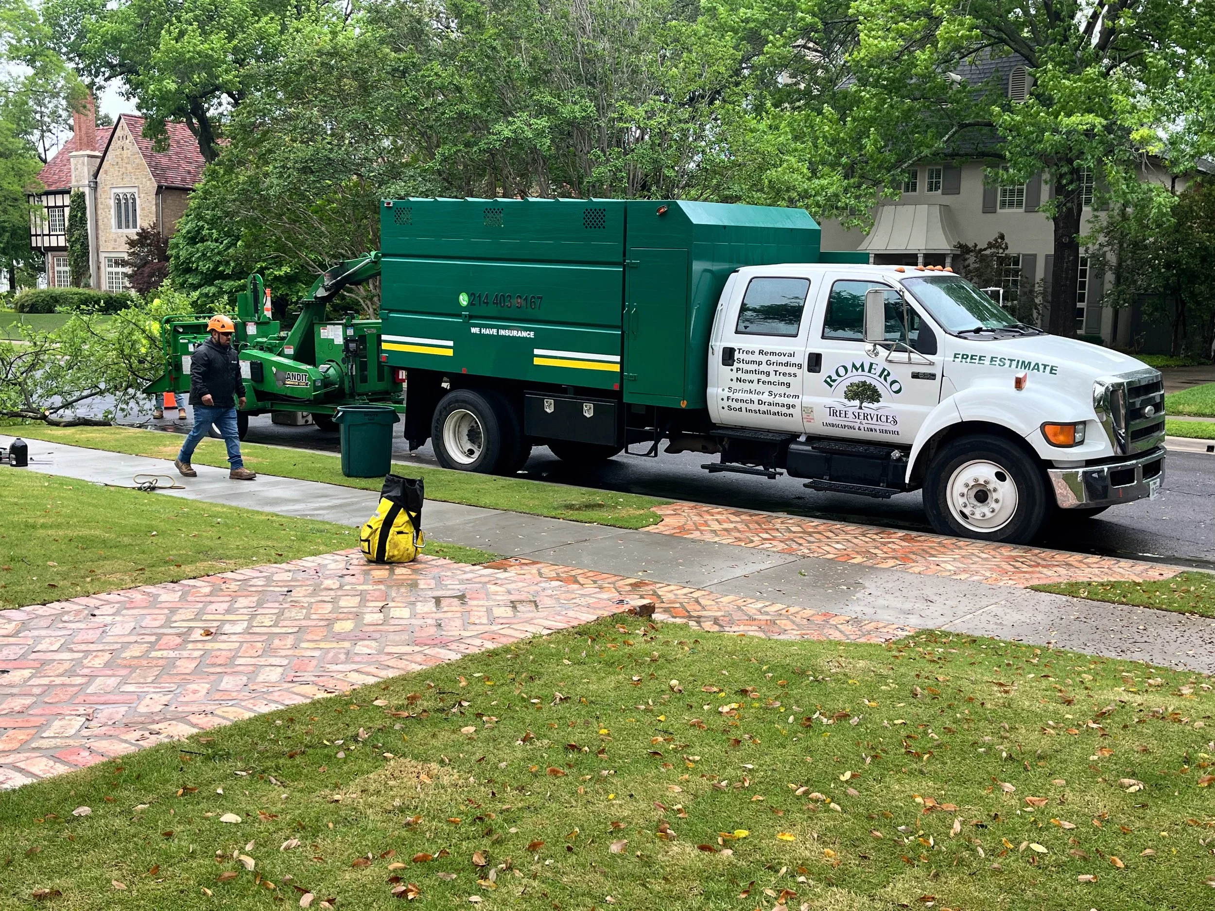 Romero Tree Service worker in a black jacket and orange helmet operating a green tree removal truck parked on a residential street. The scene is surrounded by trees and houses with lush green lawns.